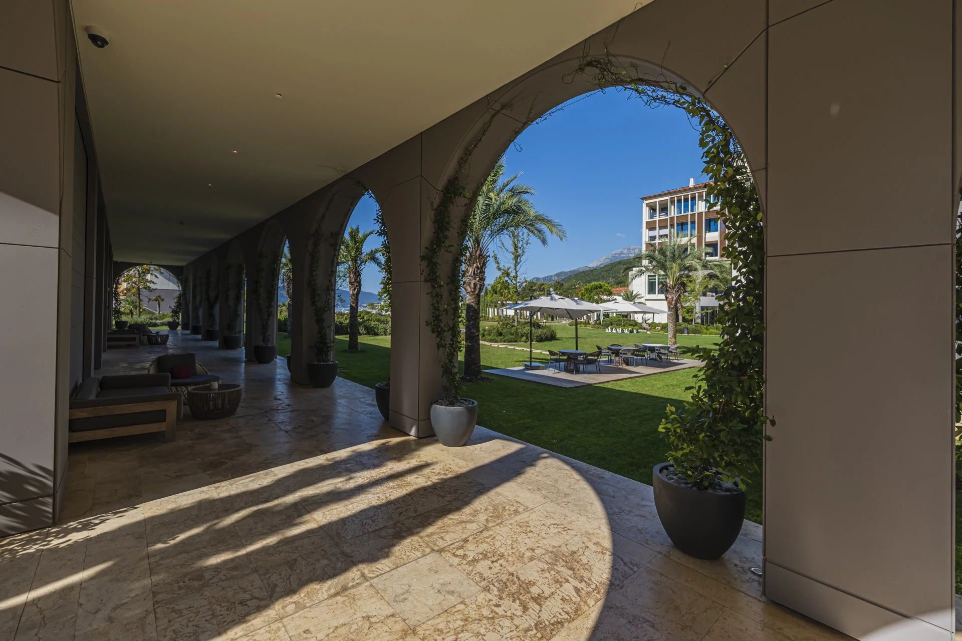 View from a covered patio overlooking a landscaped garden with palm trees, outdoor seating area with umbrellas, and a large modern building in the background with mountains beyond.