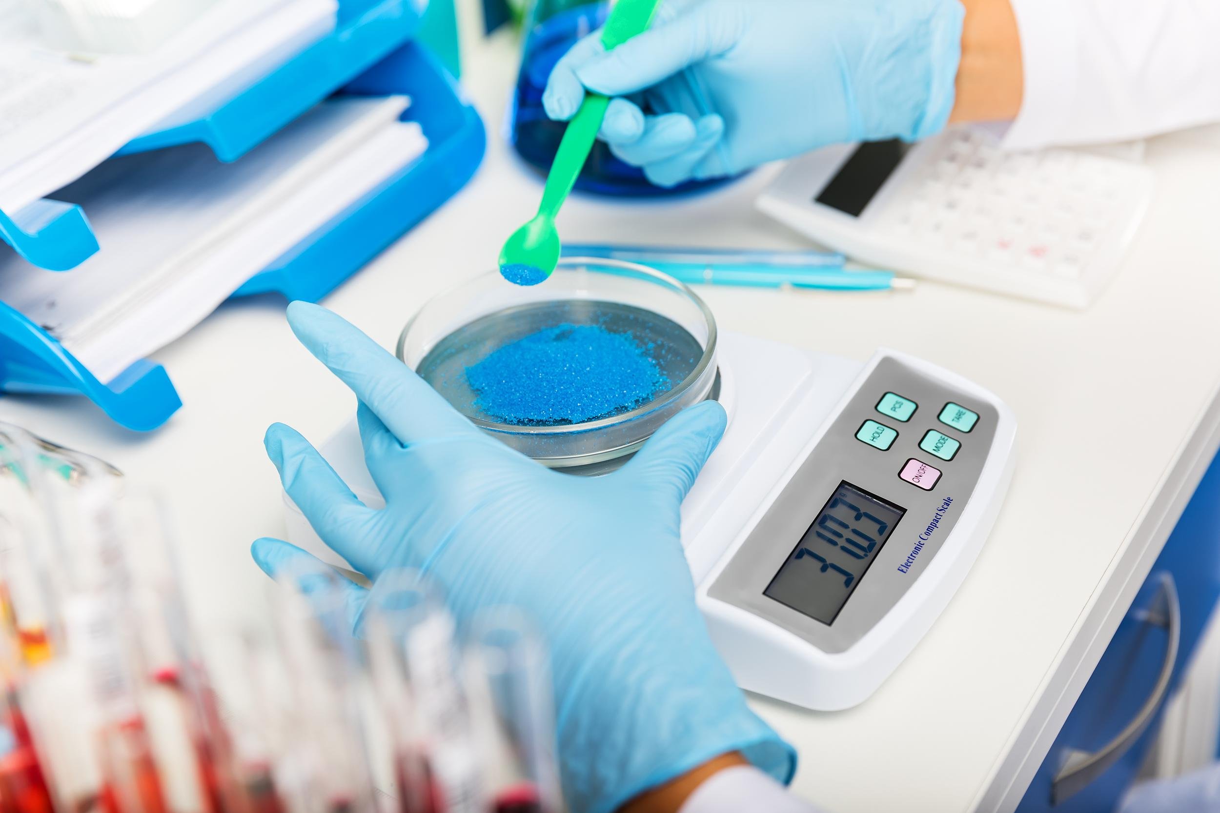 Person wearing blue gloves using a green spatula to handle blue granular substance in a clear petri dish in a laboratory setting with a digital scale.