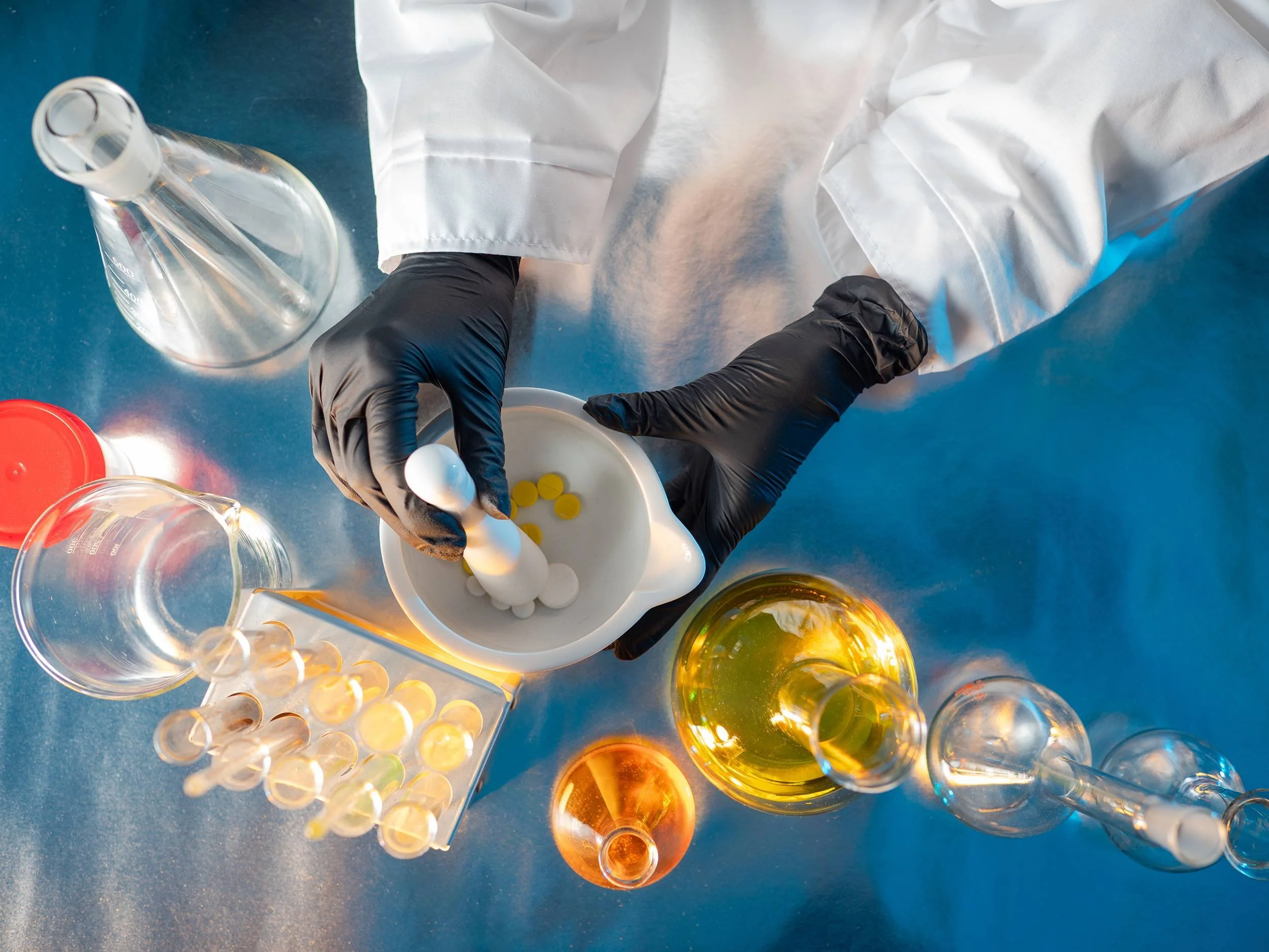 Top-down view of a lab scene with a person wearing black gloves holding a small white spoon with yellow pills, several glass containers with liquids, and a spill of white powder on a blue surface.