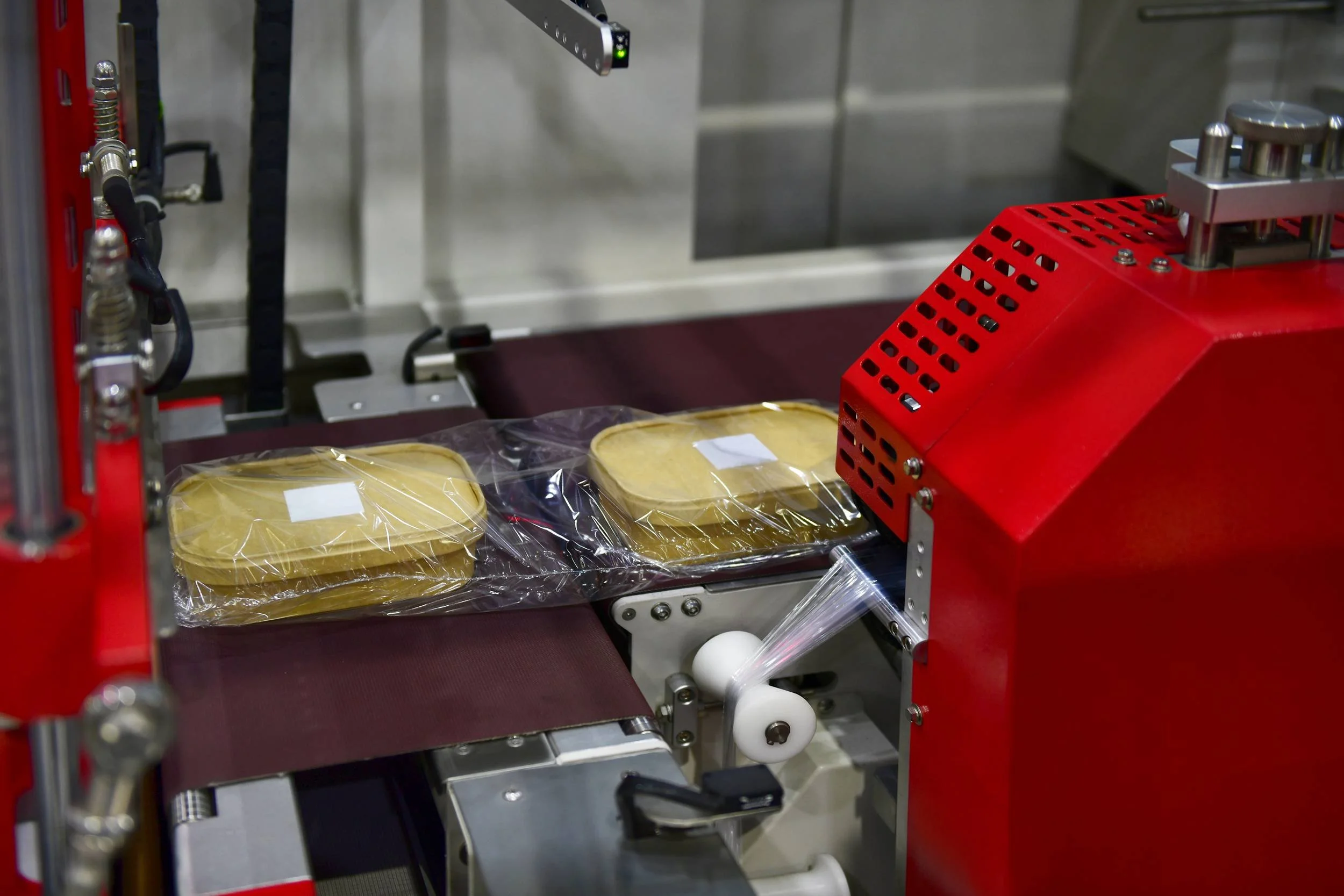 Automated packaging machine sealing plastic containers, with containers on moving conveyor belt.