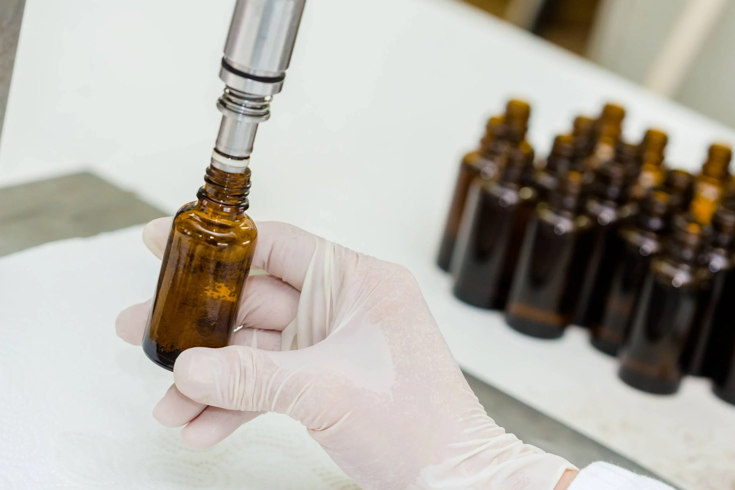 A person's hand wearing a latex glove holding a small amber glass bottle while a pen disinfects the bottle's opening. In the background, more amber bottles are arranged in a rack on a white surface.