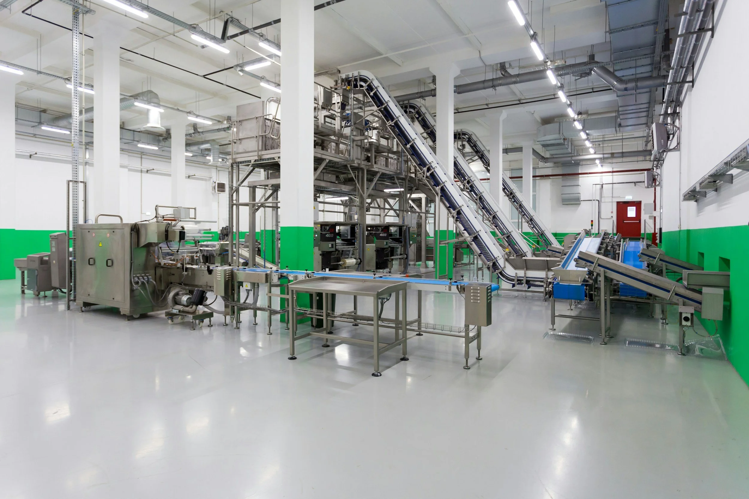Empty industrial manufacturing or processing facility with conveyor belts, stainless steel machinery, and white walls with green lower wall trim.