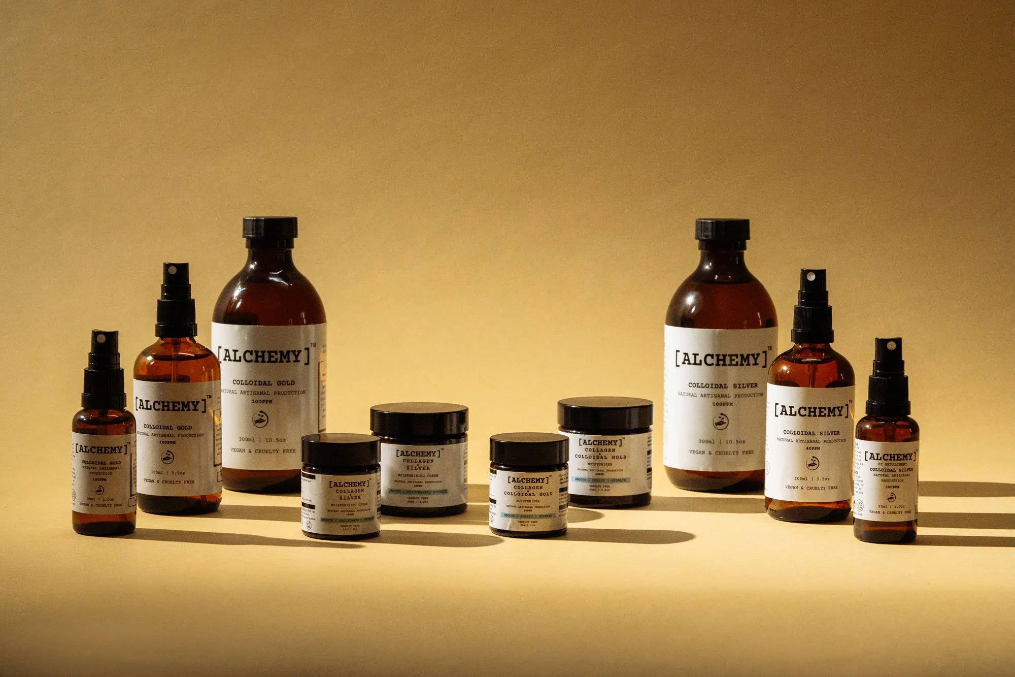 A collection of skincare products in brown bottles and jars with black caps, labeled with the brand name 'Alchemy', set against a beige background.