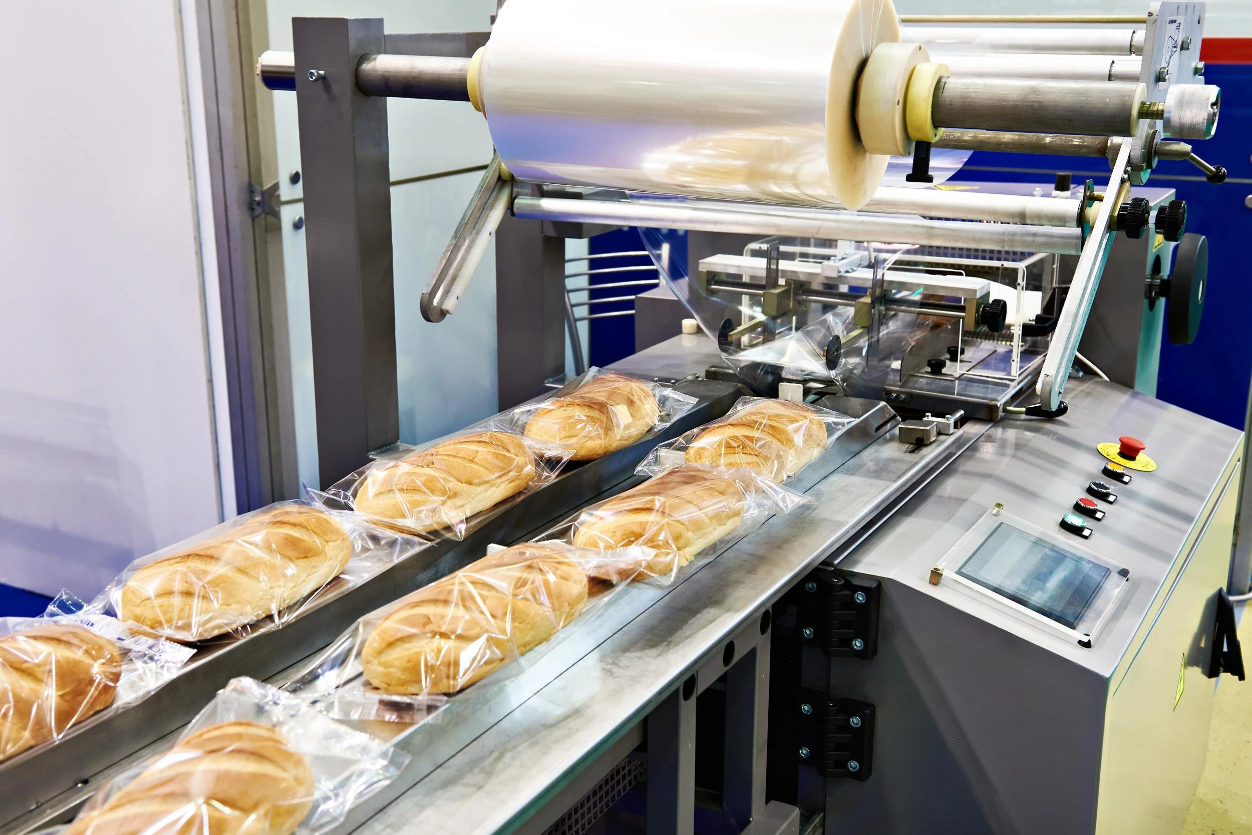 A packaging machine sealing bread rolls in plastic bags on a conveyor belt in a factory setting.