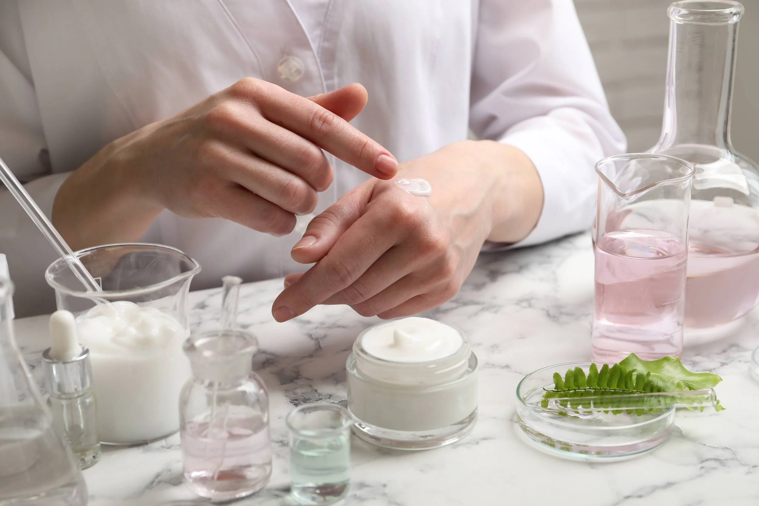 A person in a white lab coat applying a cream or lotion to their hand in a laboratory setting, with various glass containers and beakers filled with different liquids and substances on a marble countertop.