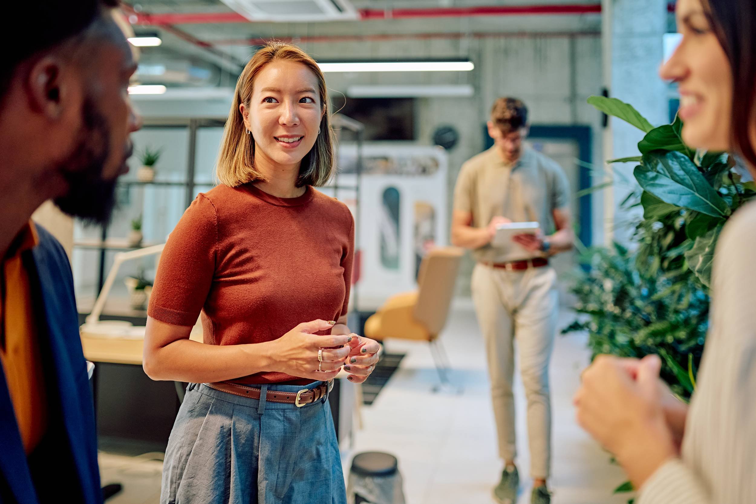 A woman in a rust-colored top and gray pants engaging in conversation with another person in an indoor office or workspace with other individuals and plants in the background.