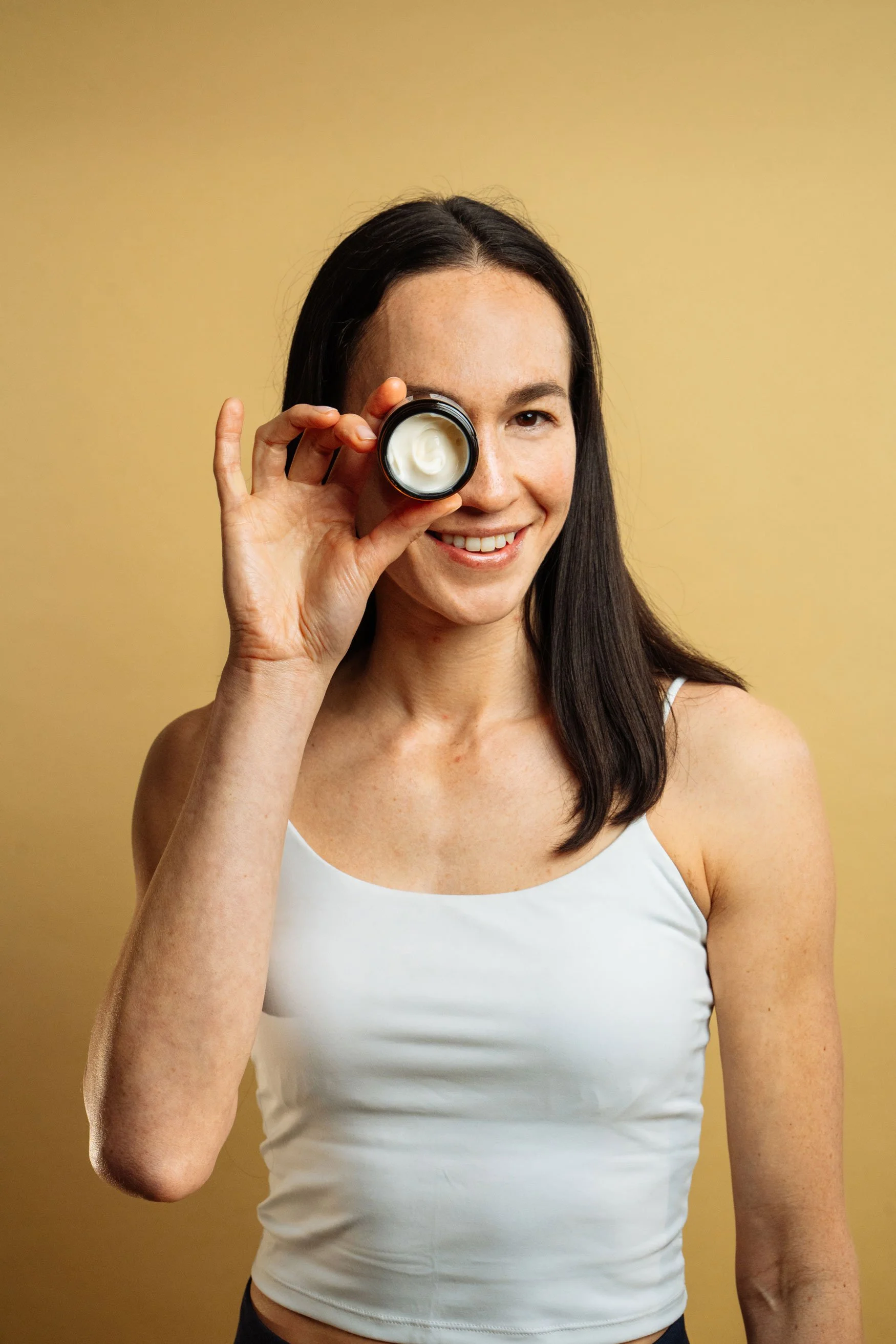 A woman with dark hair, wearing a white tank top, smiling, holding a small container of cream up to her right eye against a plain beige background.