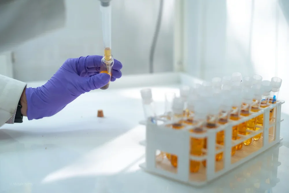 A gloved hand holds a small vial of yellow liquid in a laboratory with a rack of similar vials in the background.
