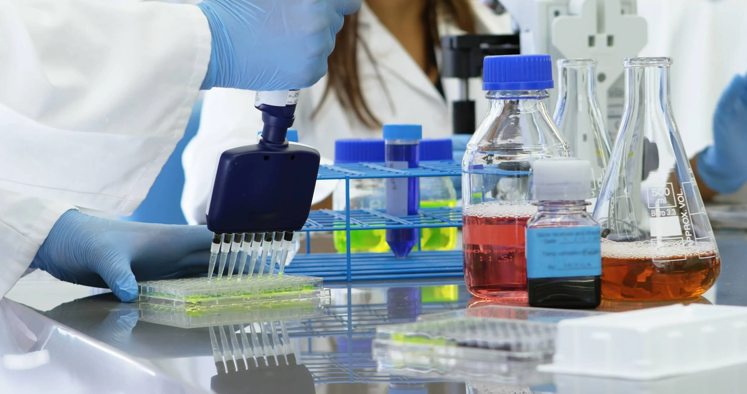 A scientist wearing blue gloves using a pipette to transfer liquid into a multi-well plate in a laboratory with various test tubes and beakers filled with colorful liquids.