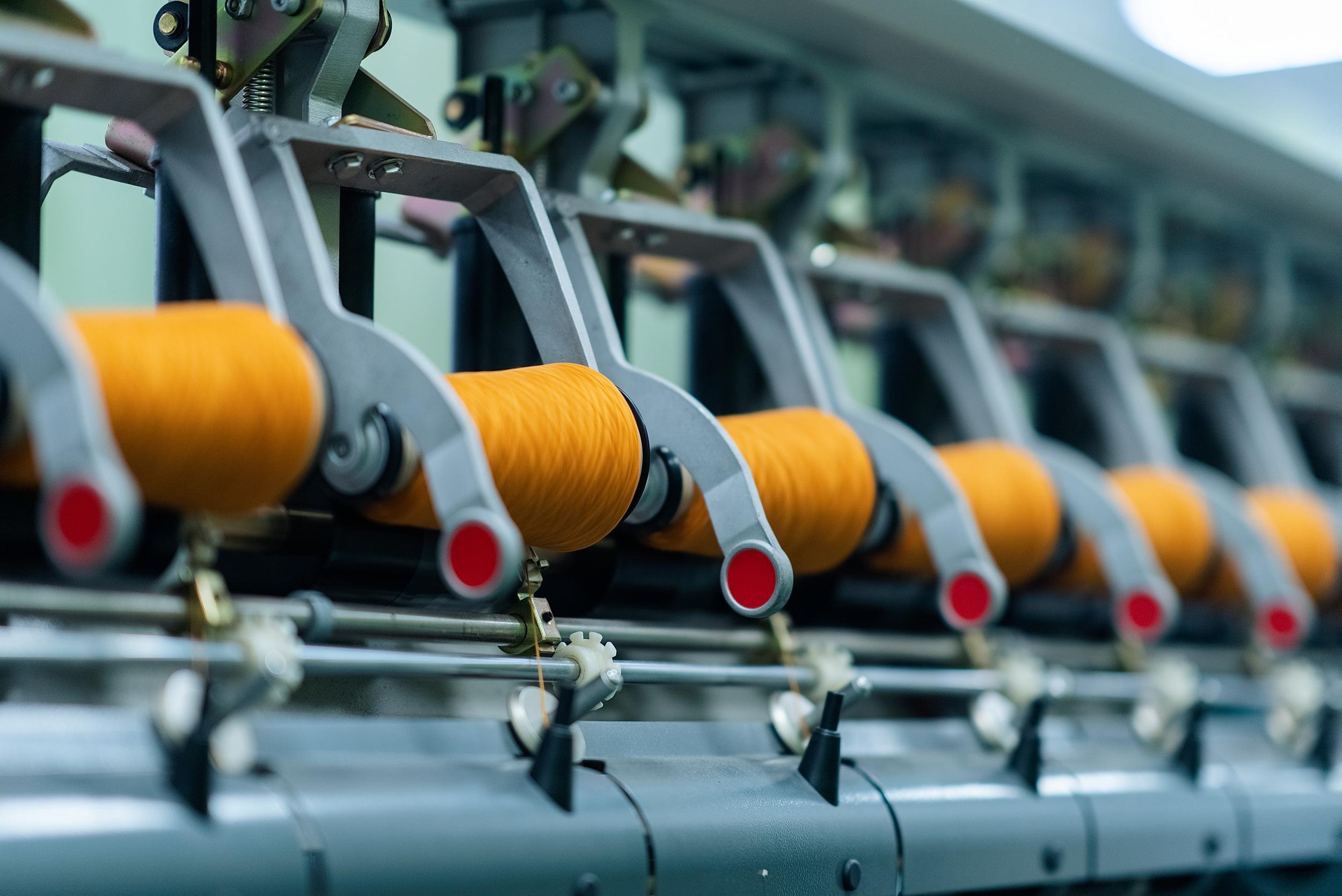 Close-up of an industrial printing machine with orange threads wound on spools, showing mechanical components and rollers.