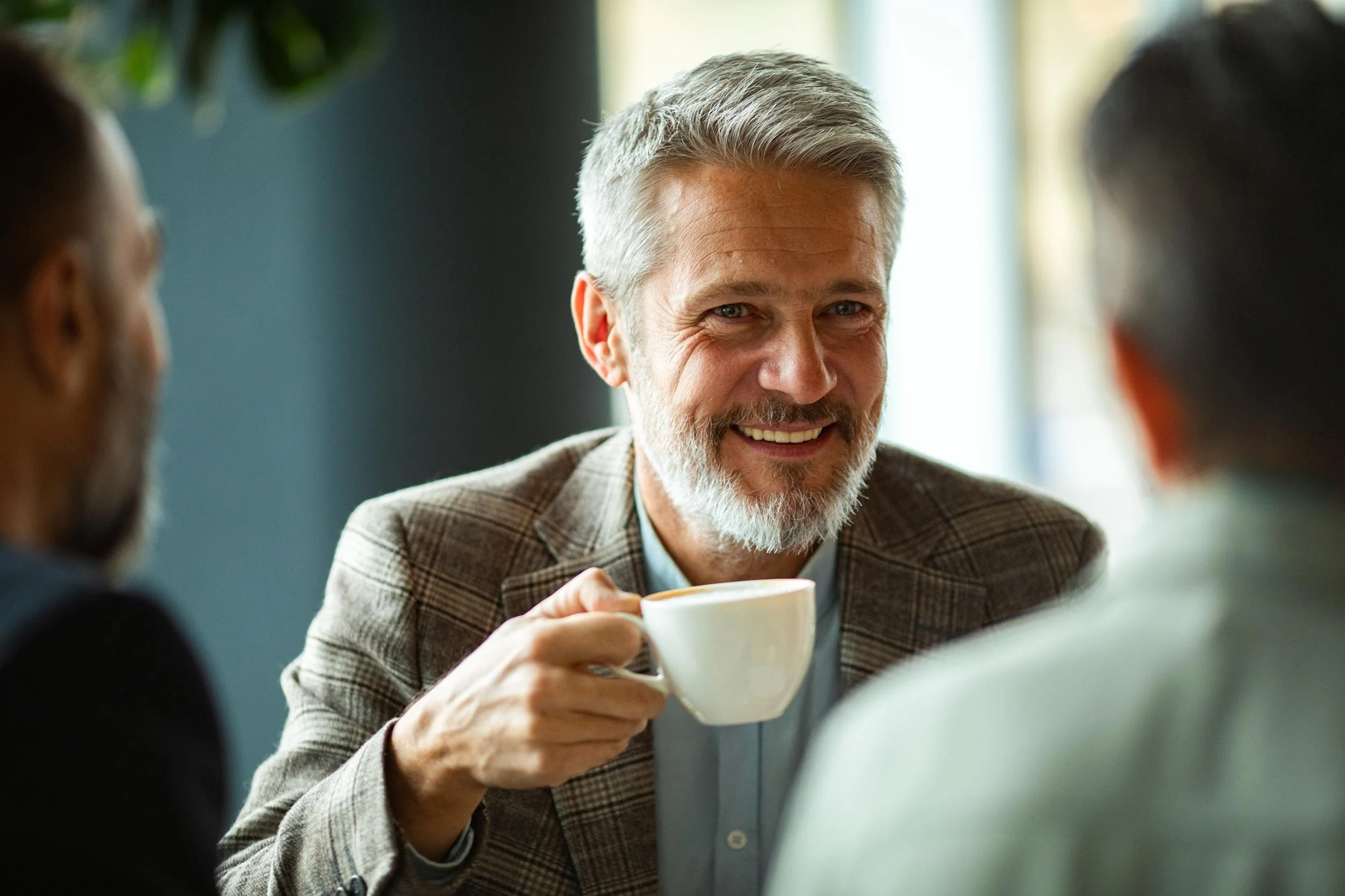 A middle-aged man with gray hair and a beard smiling while holding a coffee cup, engaging in conversation with others in a bright indoor setting.