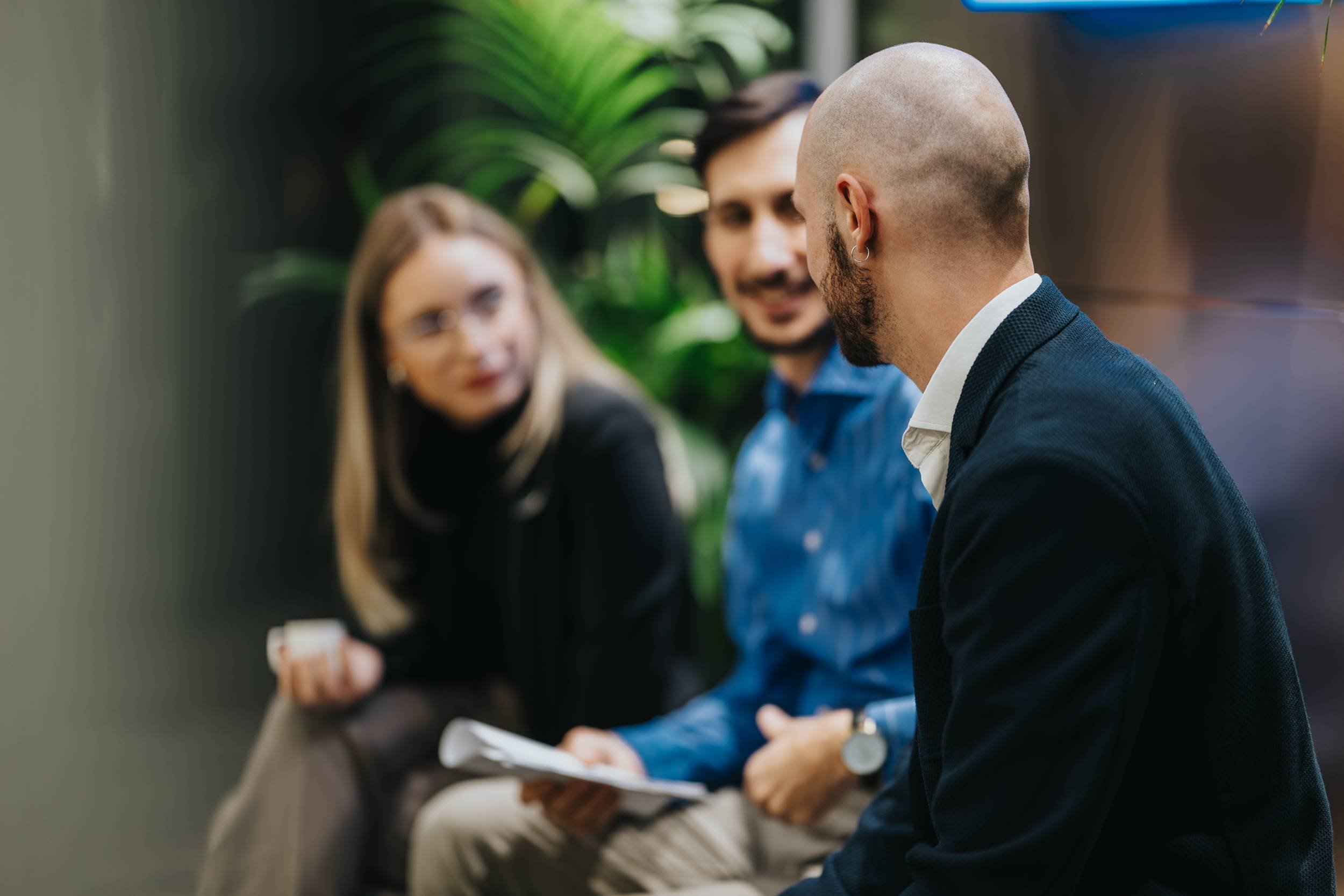 Three people sitting on a bench or sofa engaged in conversation, with a green plant in the background. The focus is on a man with a shaved head and beard wearing a dark blazer, seen from behind.