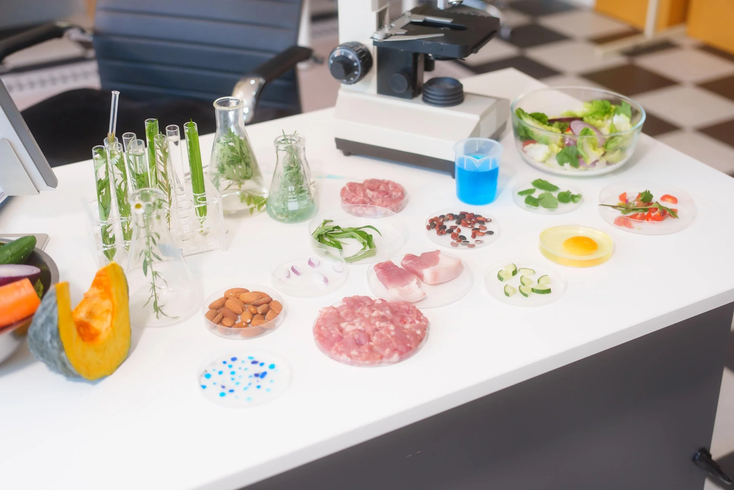 Table with laboratory equipment and food samples, including test tubes with plants, petri dishes with herbs, raw meat, and peppers, alongside other scientific materials like a microscope and a beaker with blue liquid, in a scientific or research setting.