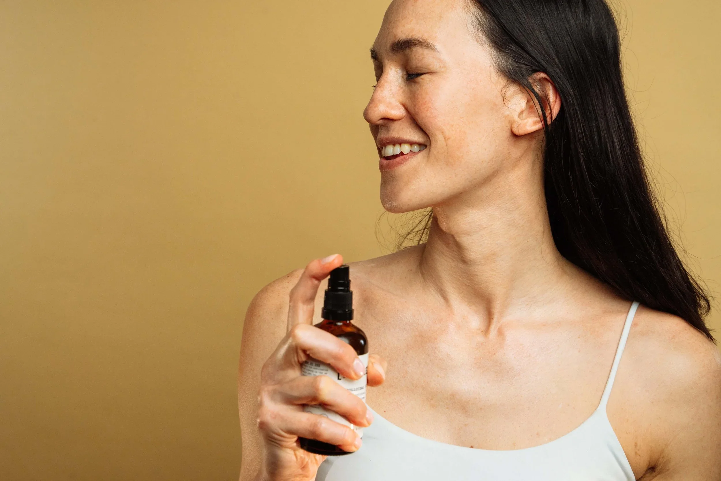A woman with long dark hair smiling with closed eyes, holding a small bottle with a dropper, wearing a white tank top, against a plain yellow background.