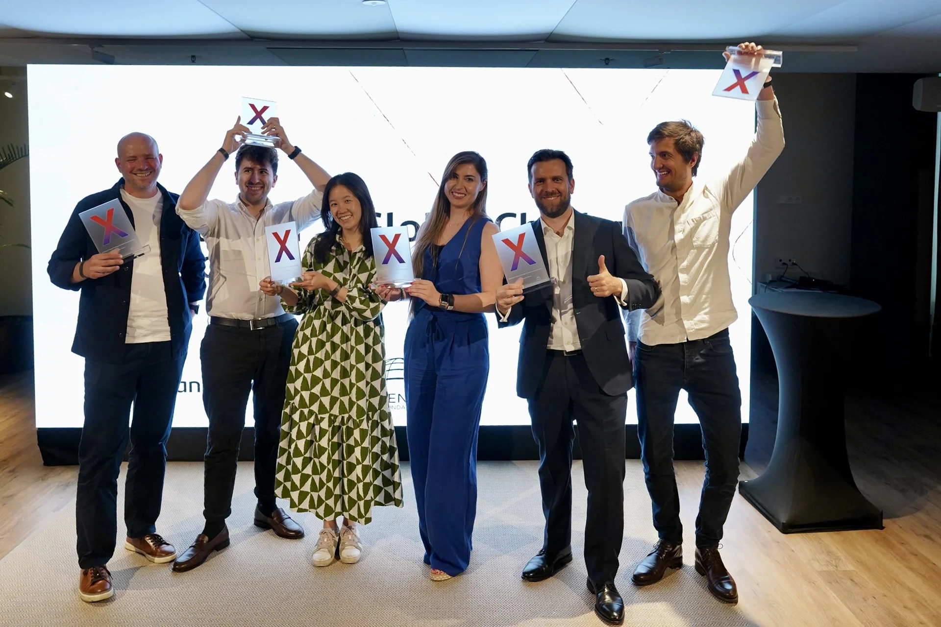 A group of seven people standing together indoors in front of a bright white screen, holding awards with a colorful X logo. They are smiling and appear celebratory, dressed in business casual or formal attire.