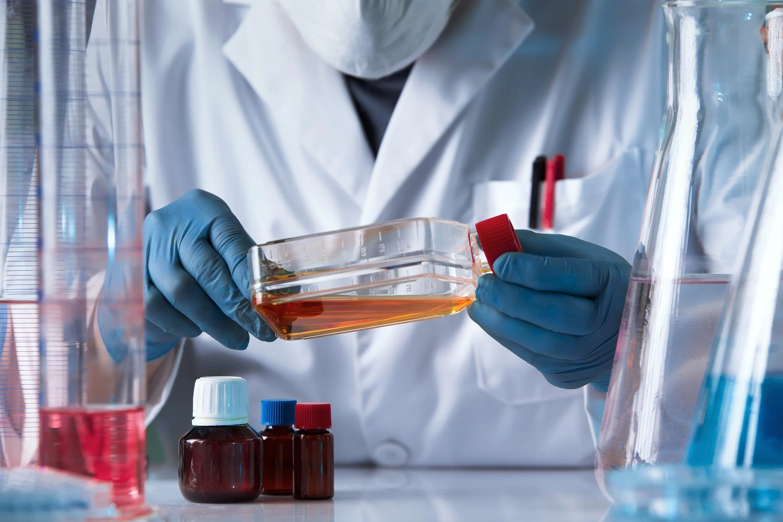 A scientist wearing blue gloves and a white lab coat is holding a petri dish with an orange liquid in a laboratory setting, surrounded by various bottles and laboratory equipment.