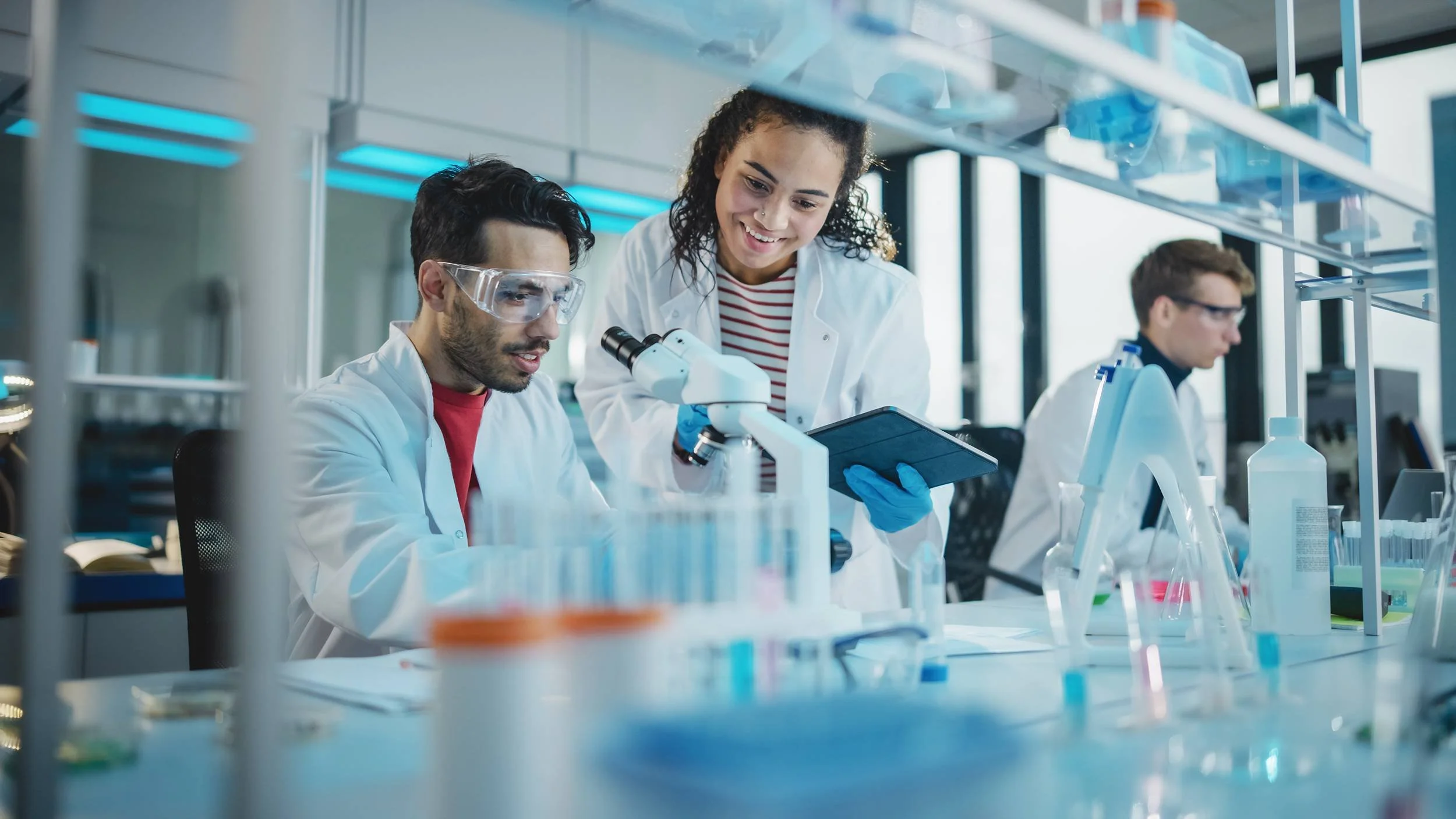Scientists in a laboratory working with microscopes, test tubes, and scientific equipment, with one scientist showing a tablet to another.