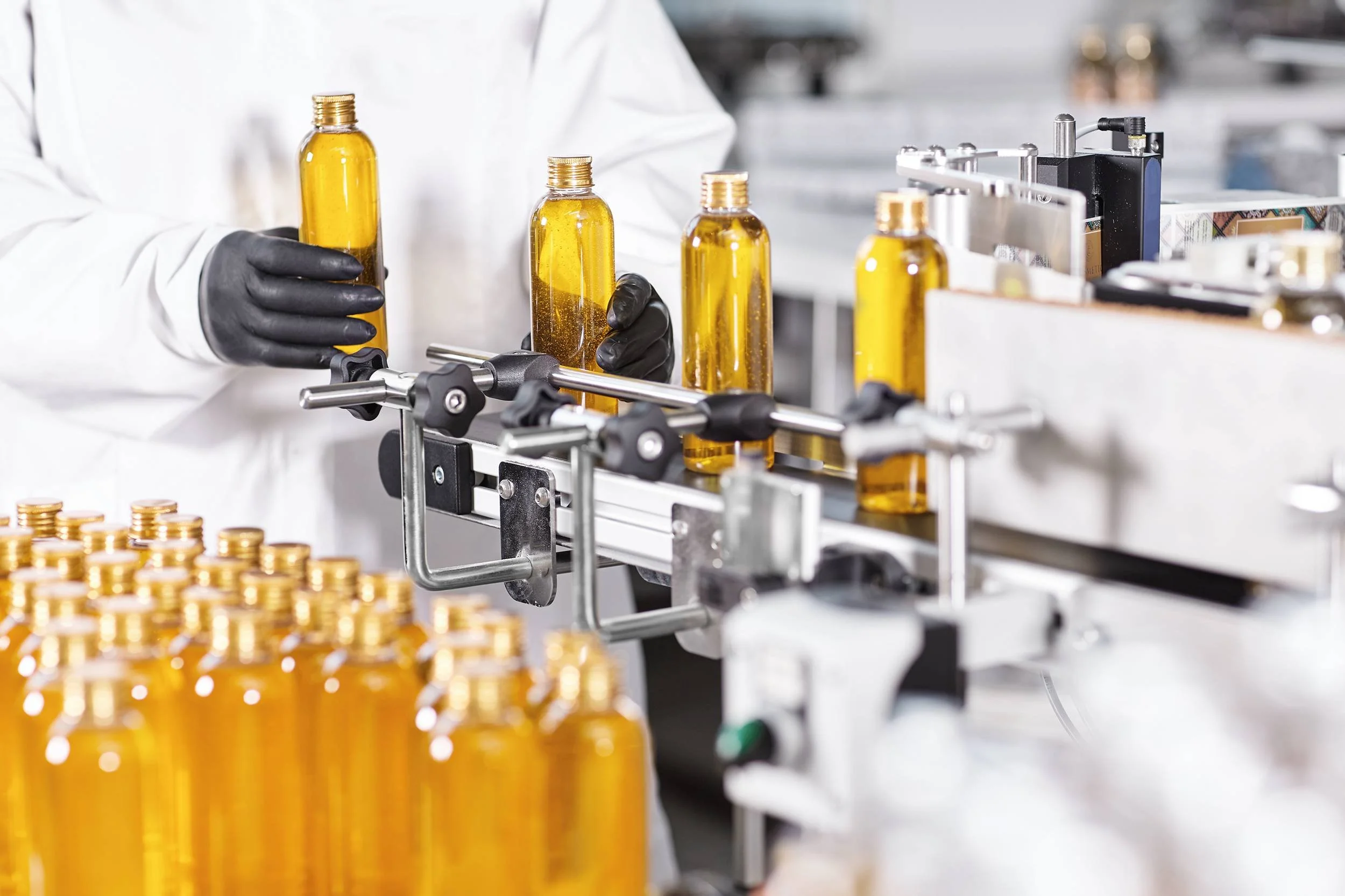 A worker in a white lab coat and black gloves assembles glass bottles on a manufacturing line. The bottles are amber-colored and are being filled or capped as they move along the conveyor belt. Some empty bottles and small stacks of coins are visible in the foreground.