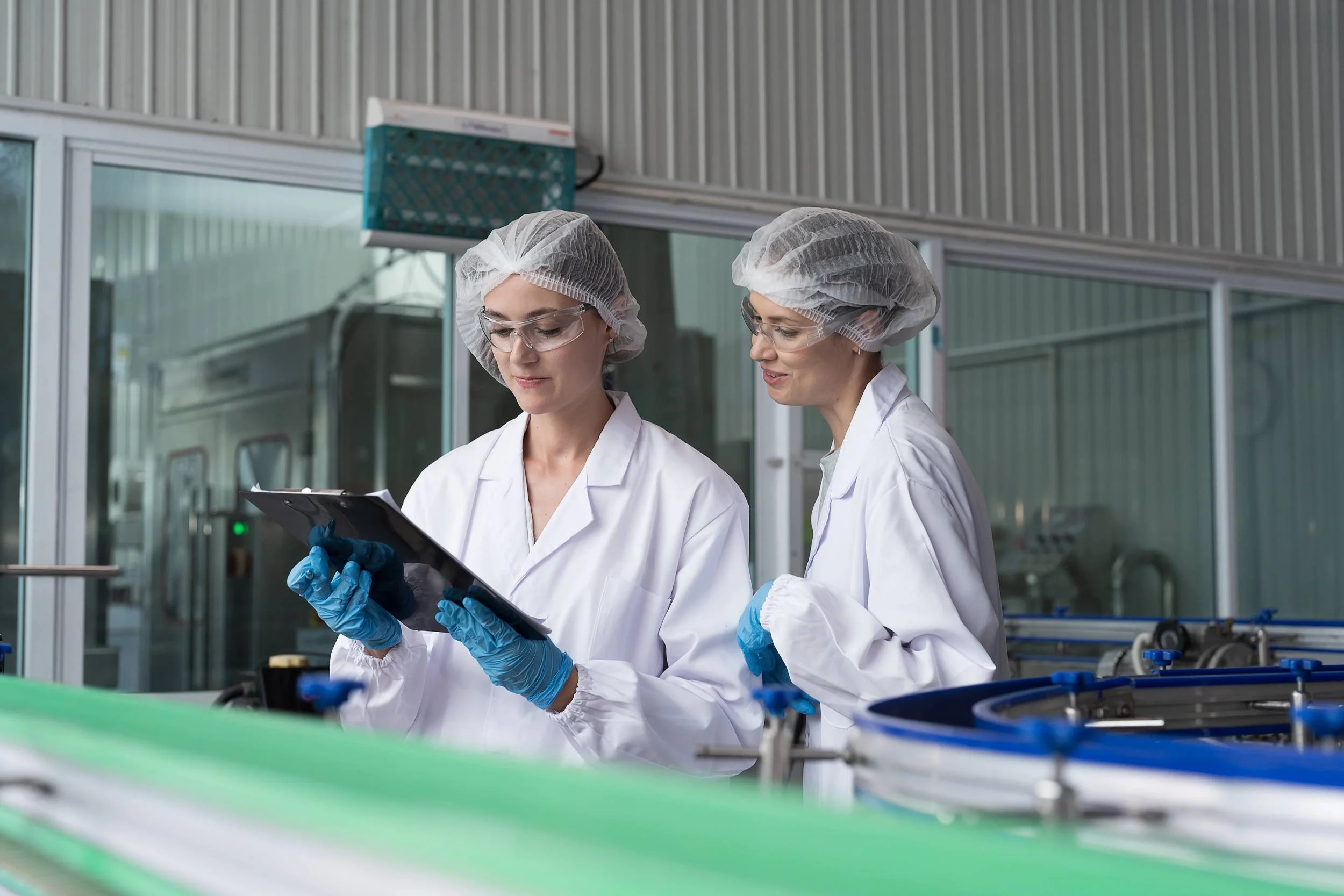 Two women wearing hairnets, lab coats, safety glasses, and gloves working in an industrial production facility.