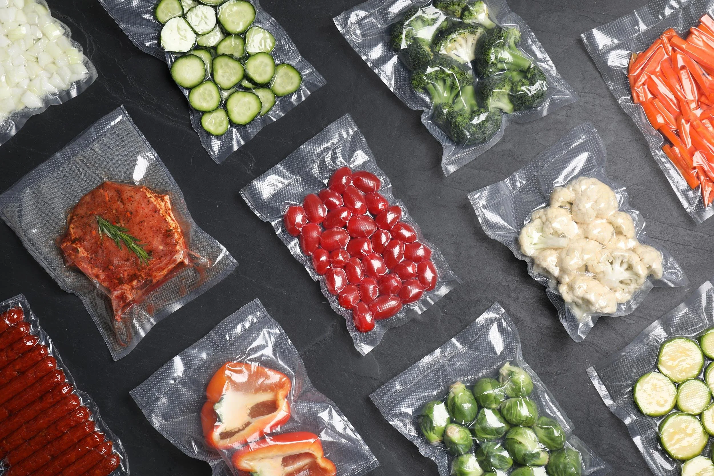Various fresh vegetables vacuum-sealed in clear plastic packaging on a dark surface, including sliced cucumbers, broccoli, cherry tomatoes, cauliflower, red bell peppers, and garlic cloves.