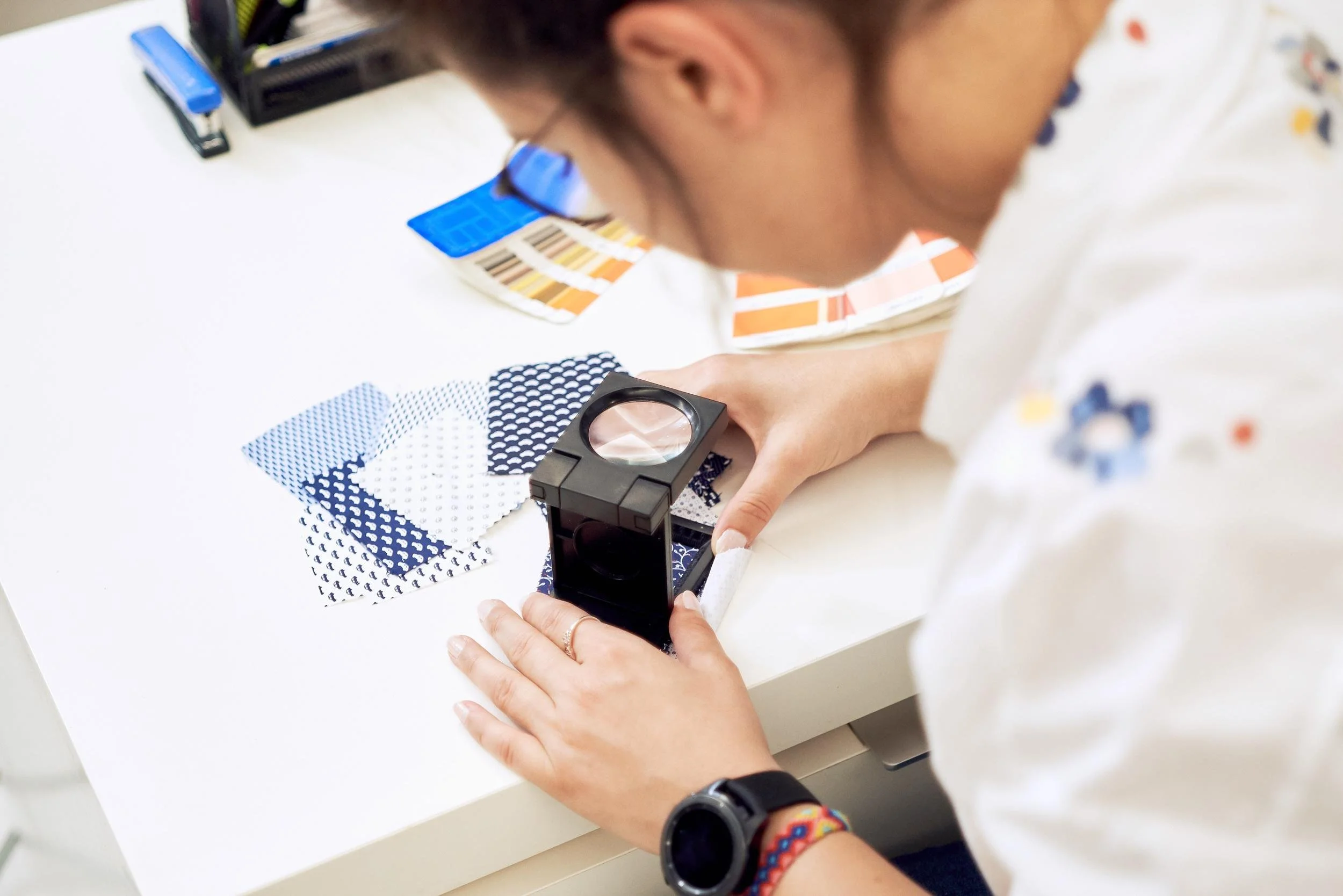 Person examining fabric samples with a magnifying glass on a white table, surrounded by color swatches and office supplies.