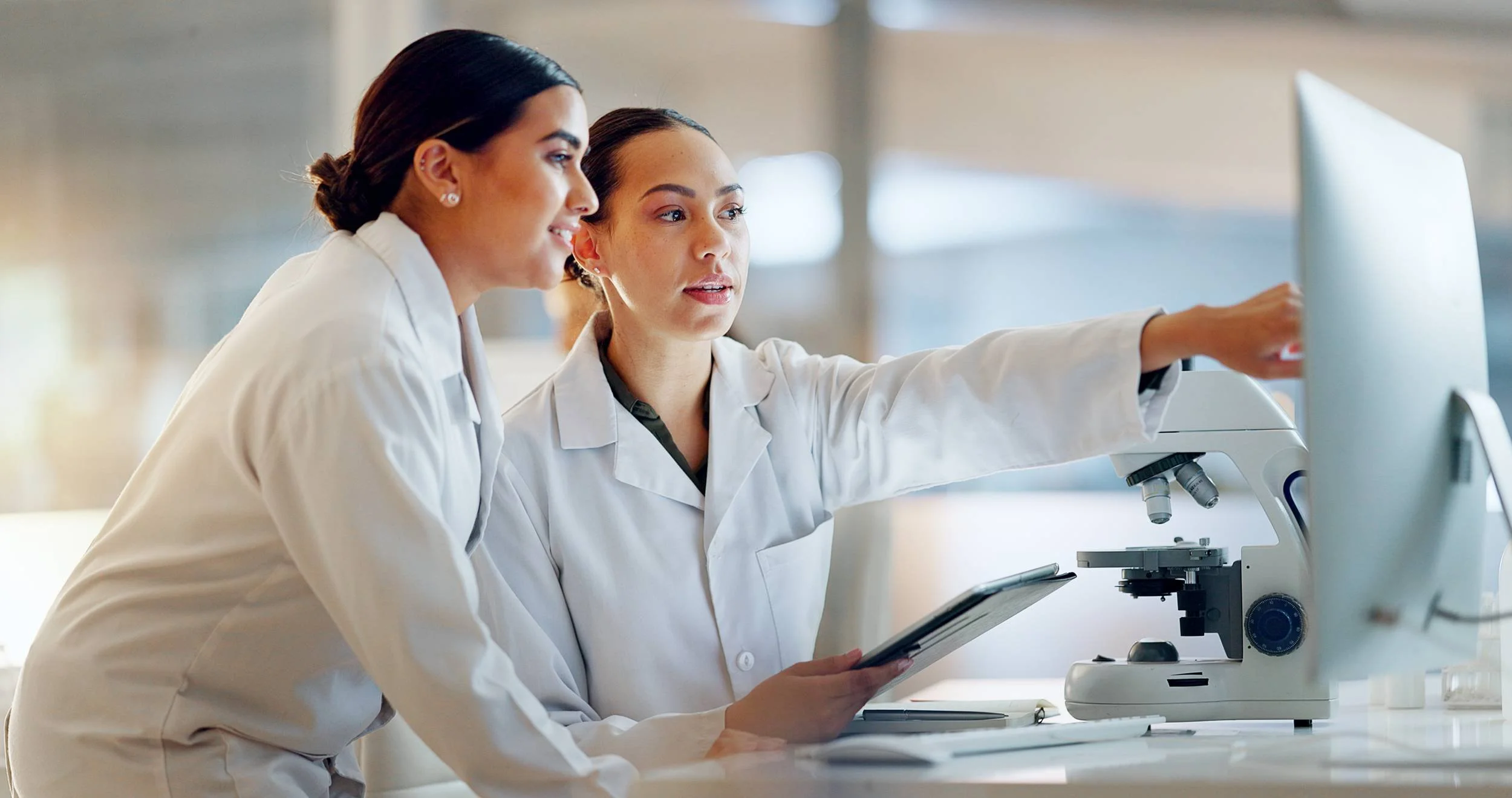 Two female scientists in lab coats working together, one pointing at a computer monitor, with a microscope on the table.