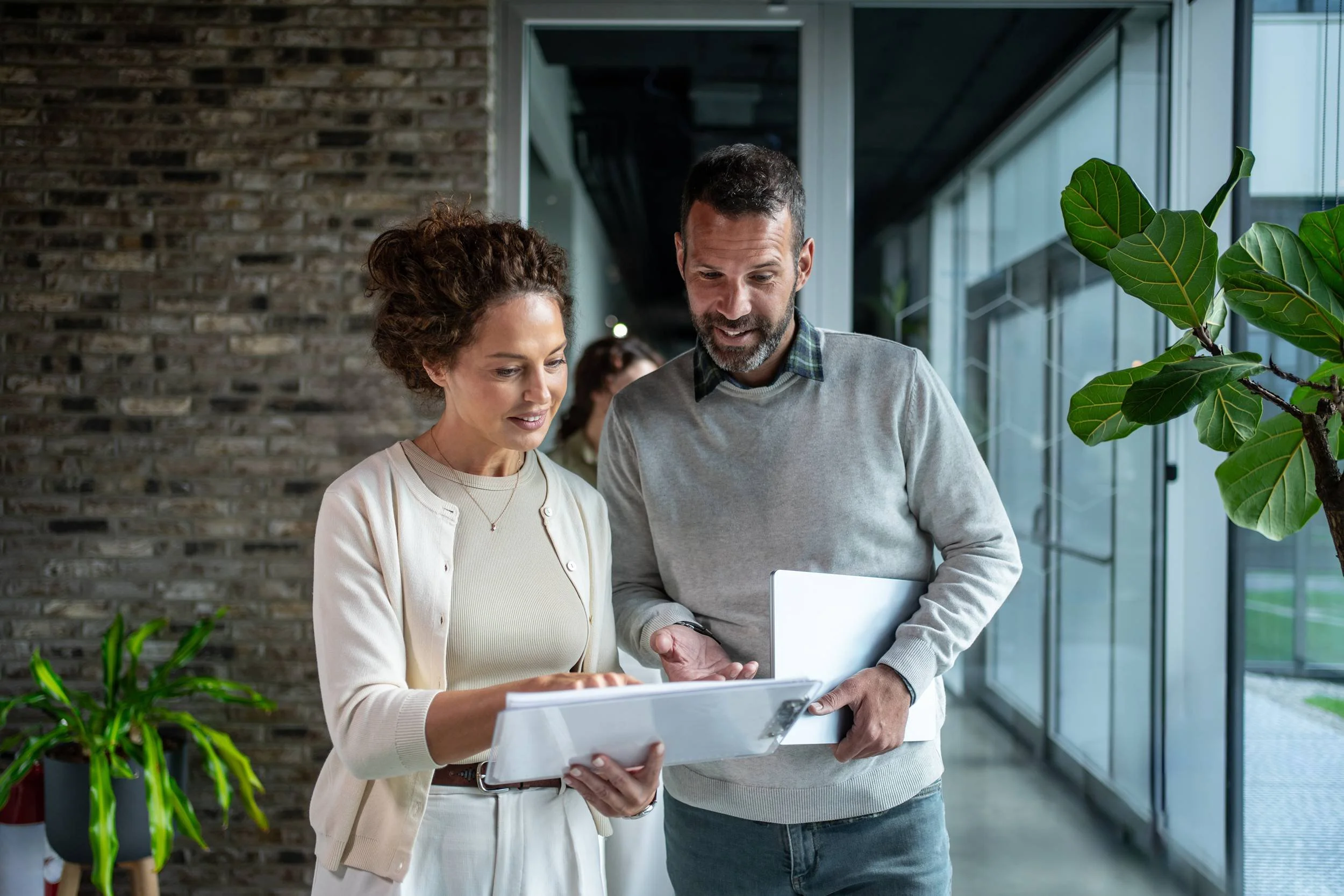 Two people, a woman and a man, are looking at a tablet while standing in a modern office space with large windows, a brick wall, and green plants.