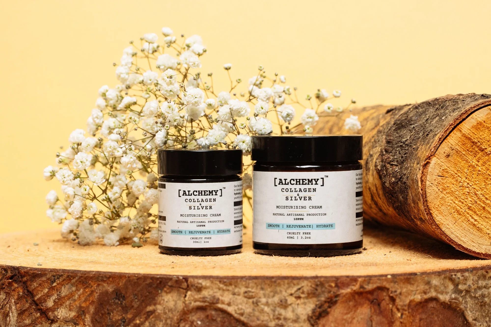 Two black jars labeled 'Alchemy Collagen & Silver Moisturising Cream' with white flowers, a wooden log, and a tree trunk cross-section on a wooden surface against a yellow background.