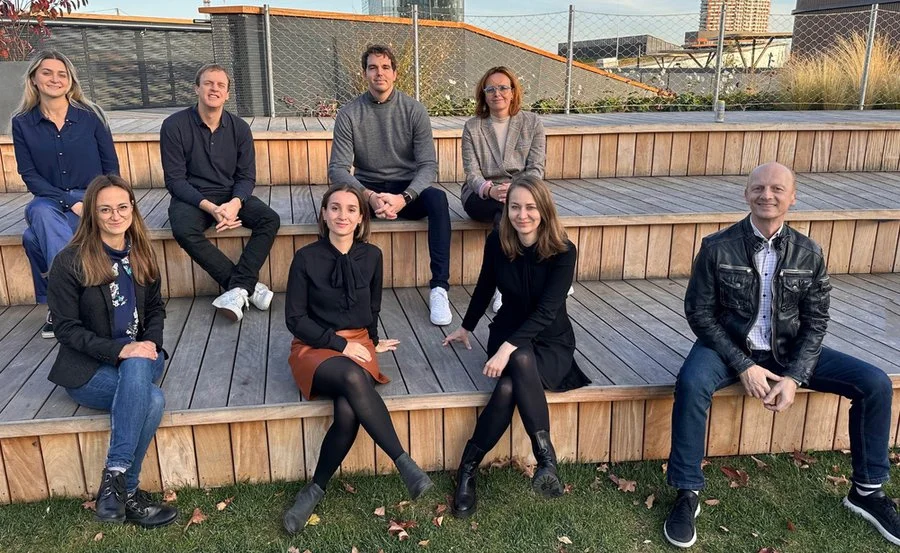 Seven people are sitting on wooden tiered seating outdoors, with a background of a fence, buildings, and grass. They are dressed casually and smiling at the camera.