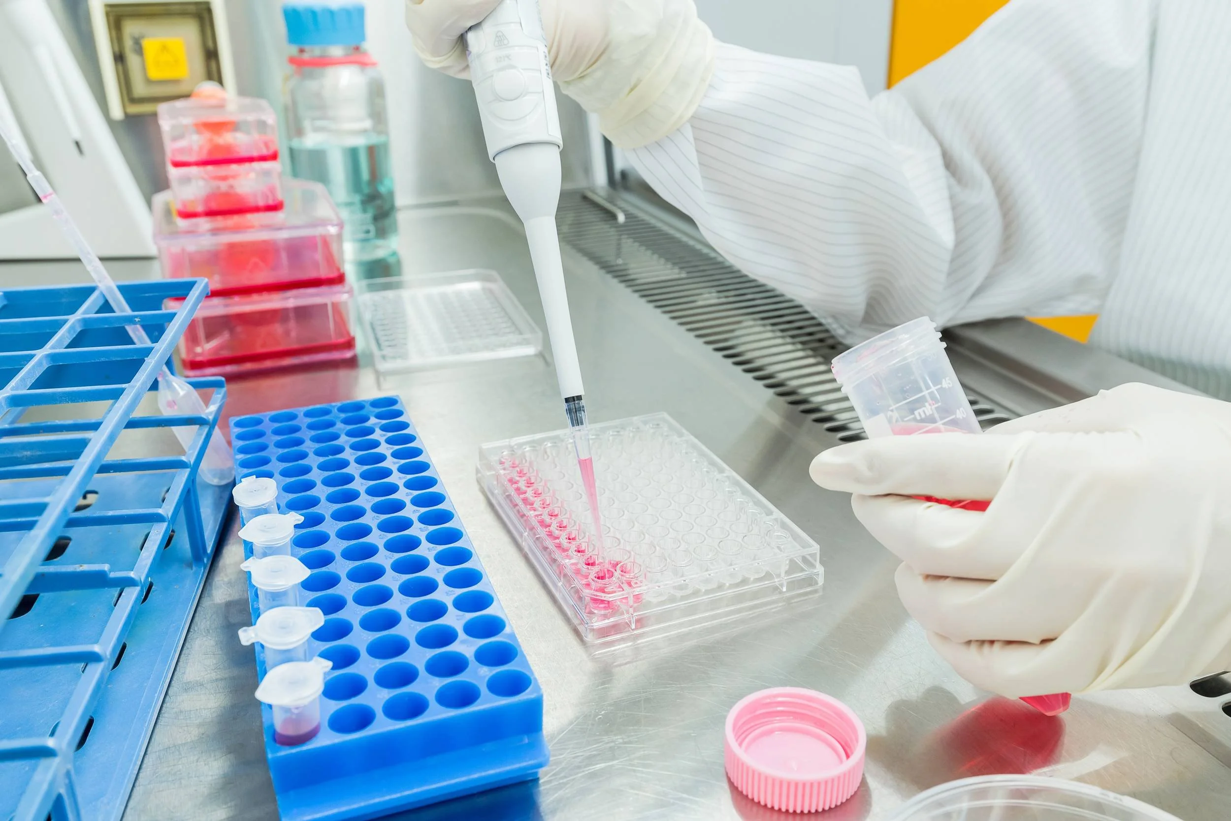 A scientist working in a laboratory, using a pipette to transfer liquid into a plastic well plate, with various laboratory supplies and containers around.