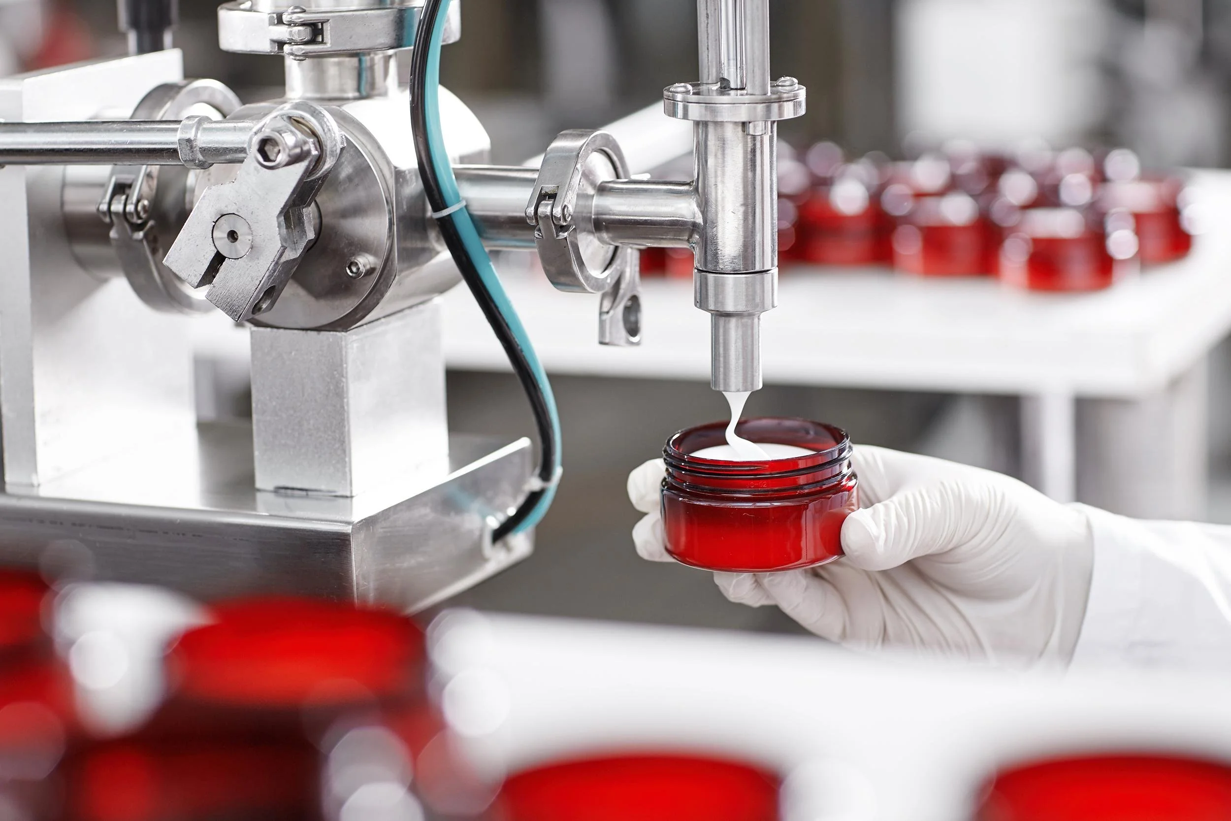 A gloved hand filling a red jar with white liquid from a machine in a laboratory or manufacturing setting.