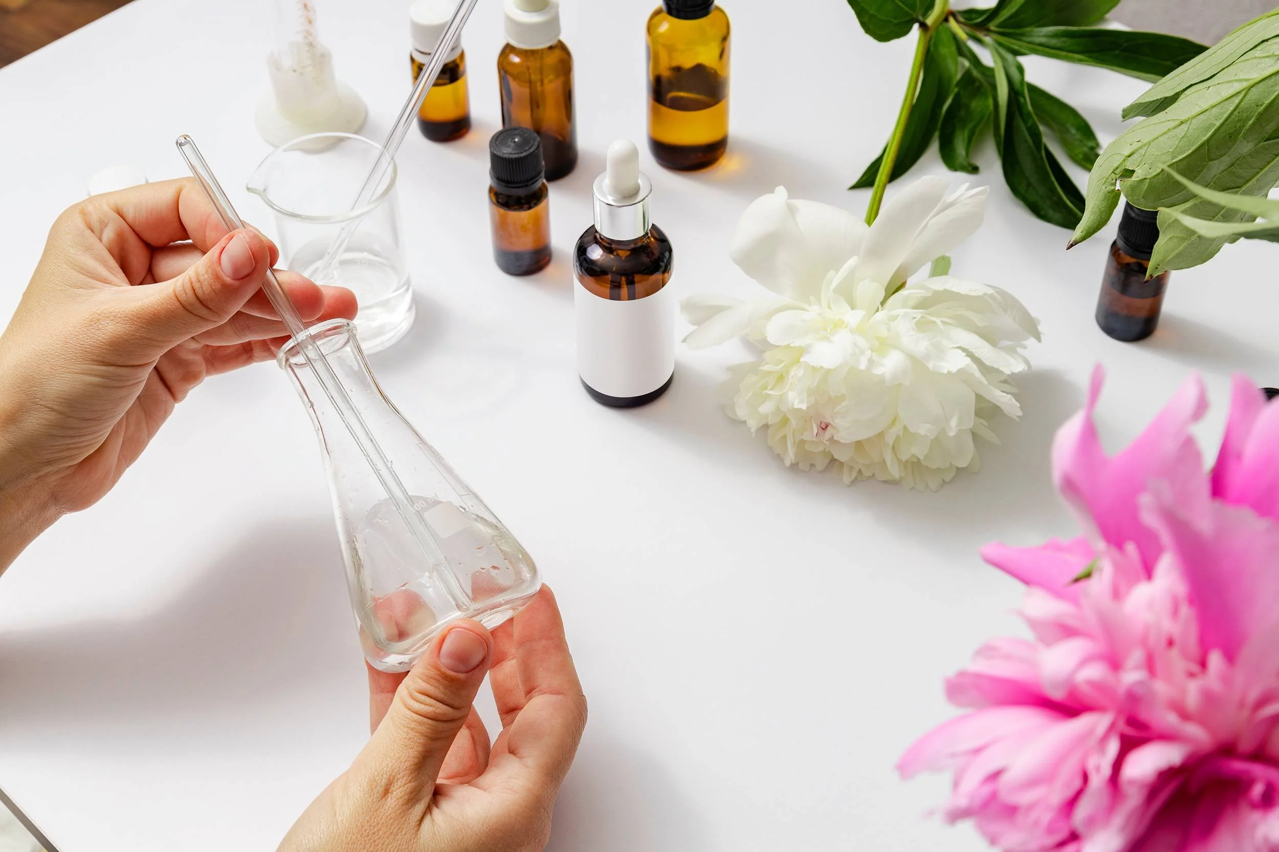 Person holding a glass dropper with a liquid, surrounded by small brown bottles, flowers, and green leaves on a white surface.
