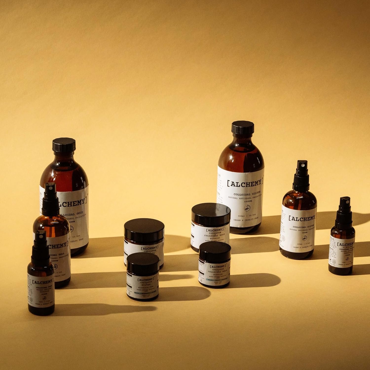 Several bottles and jars of skincare products labeled 'Alchemy,' positioned on a yellow surface with yellow background, casting shadows.