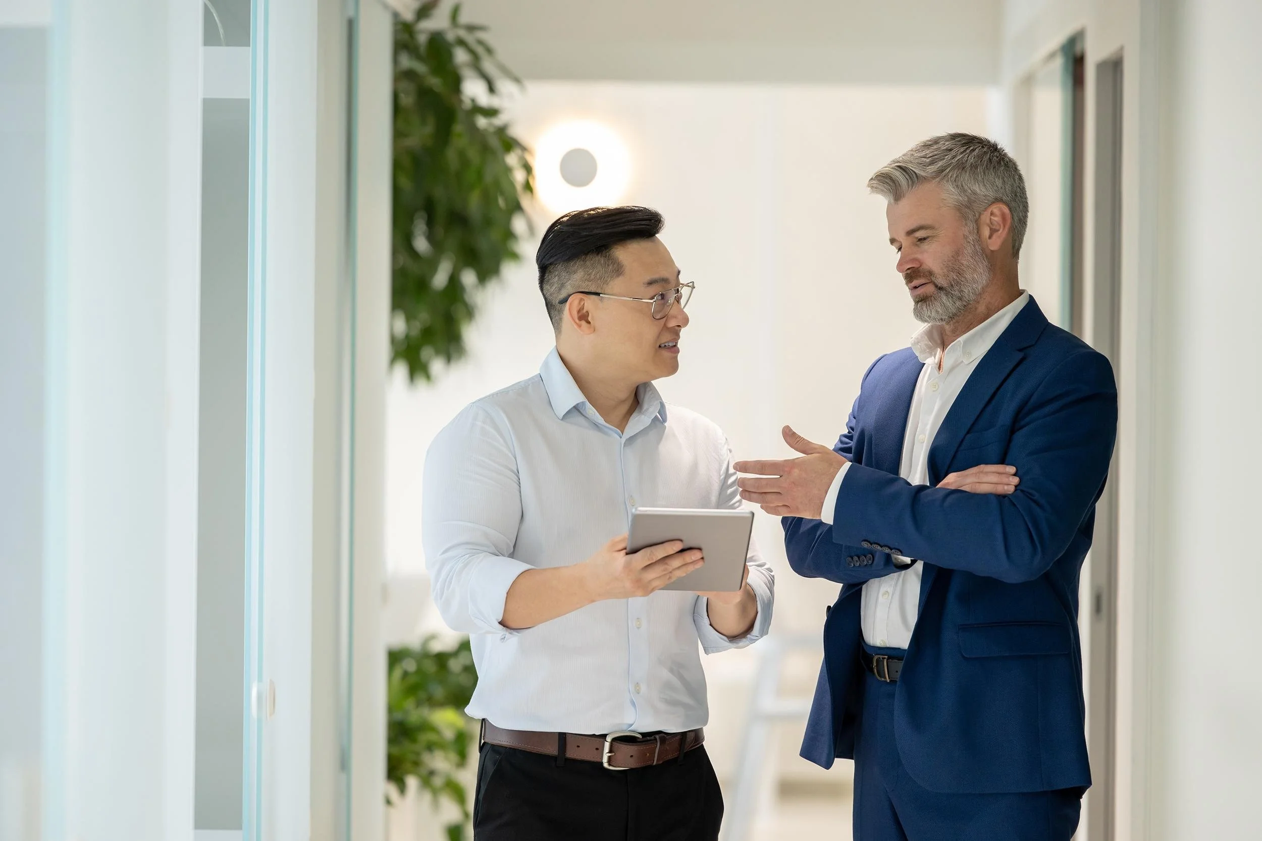 Two men, one holding a tablet, engaged in conversation in a bright, modern office hallway with white walls and green plants in the background.