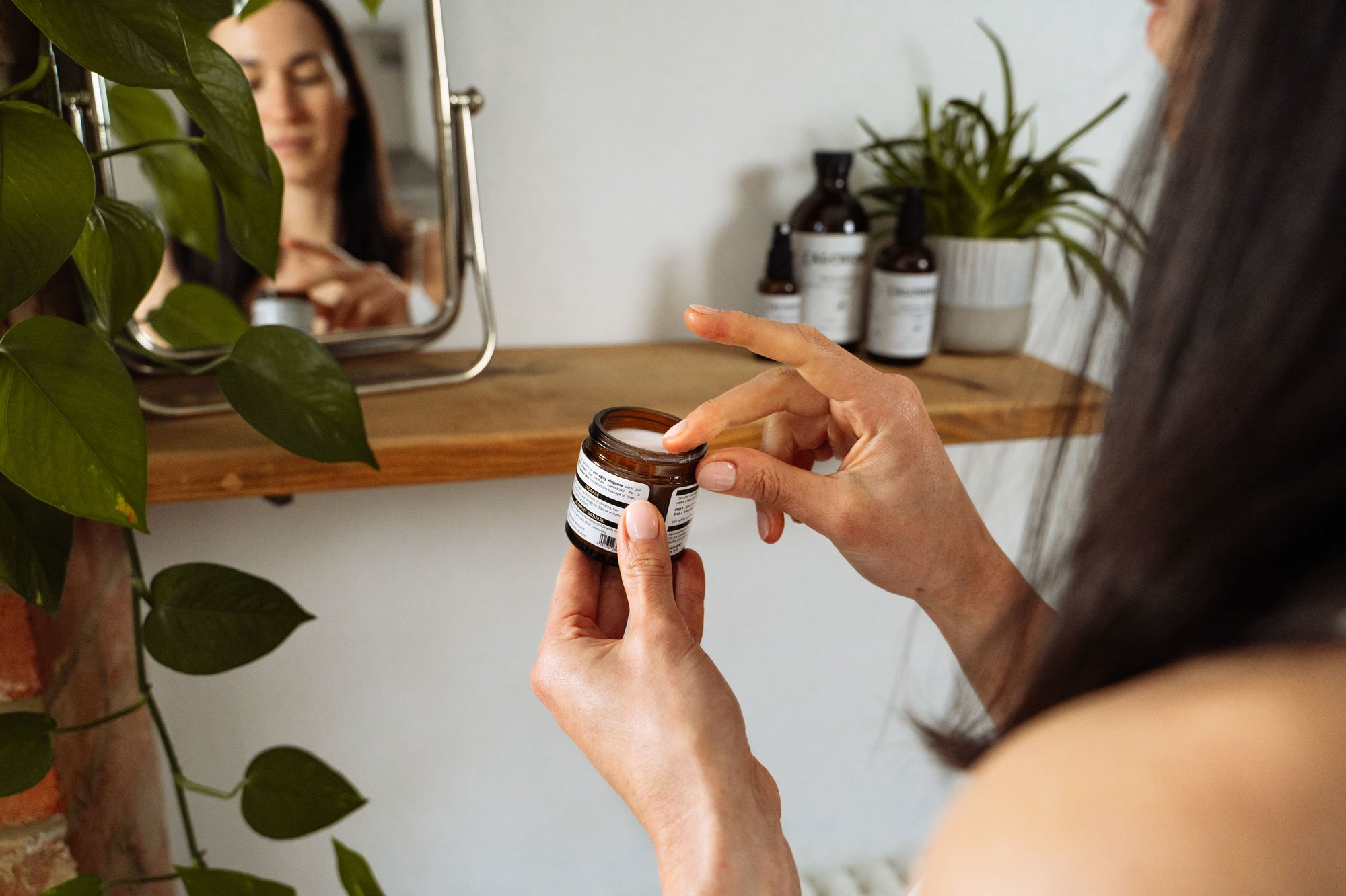Person holding an open bottle of supplement or medication, with a mirror reflecting their face, and green houseplants and bottles on a shelf in the background.