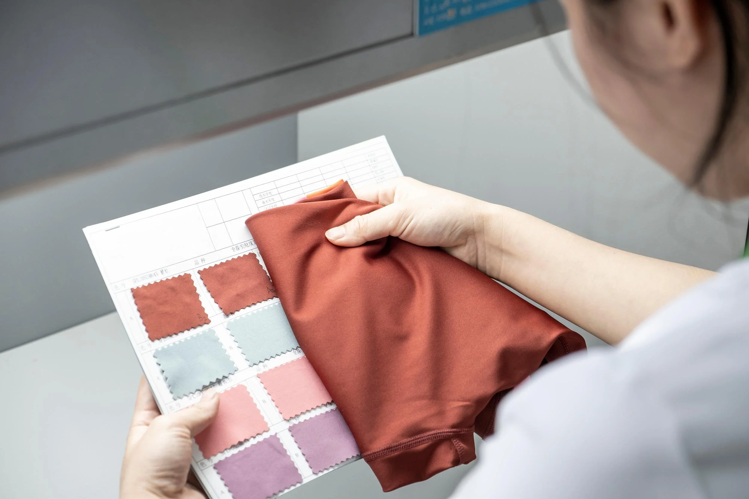 Person holding fabric color swatch booklet and testing a reddish-brown fabric with a white piece of paper in the background.