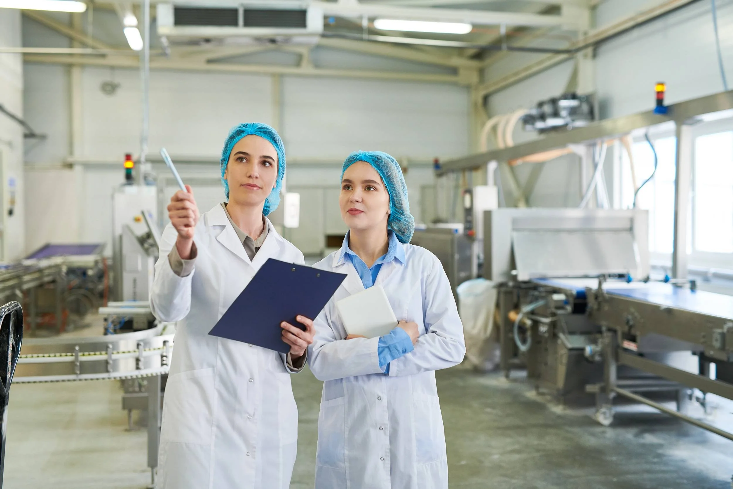 Two women in lab coats and blue hair caps in a laboratory or manufacturing facility, one holding a clipboard and pen, with equipment and machinery in the background.