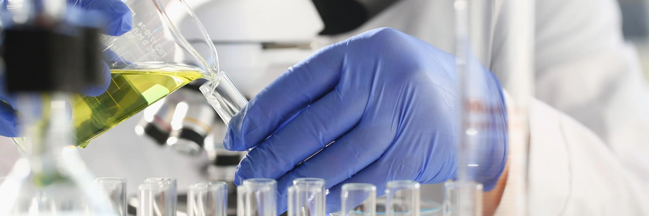 A scientist wearing blue gloves pouring a yellow-green liquid into a test tube in a laboratory.