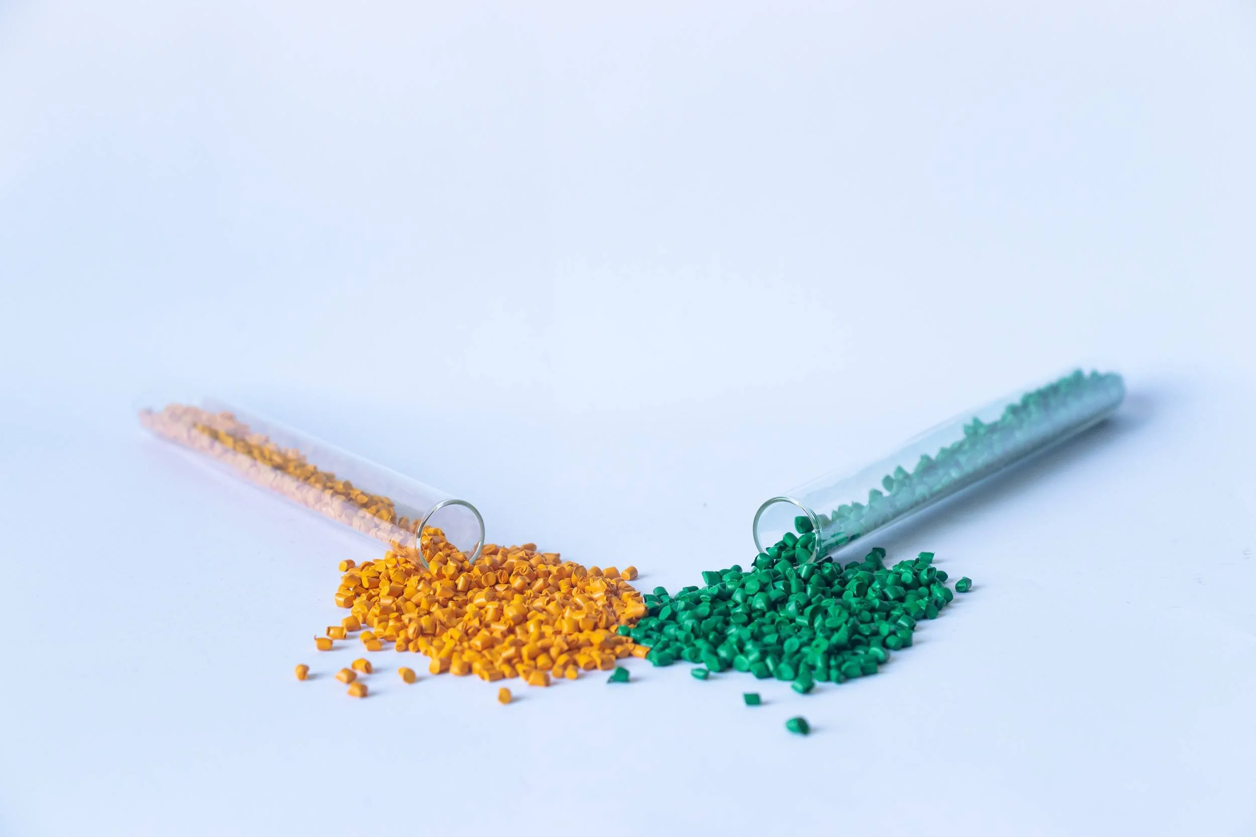 Two glass test tubes tilted on a white surface. The left test tube has orange granules spilling out, and the right test tube contains green granules spilling out.