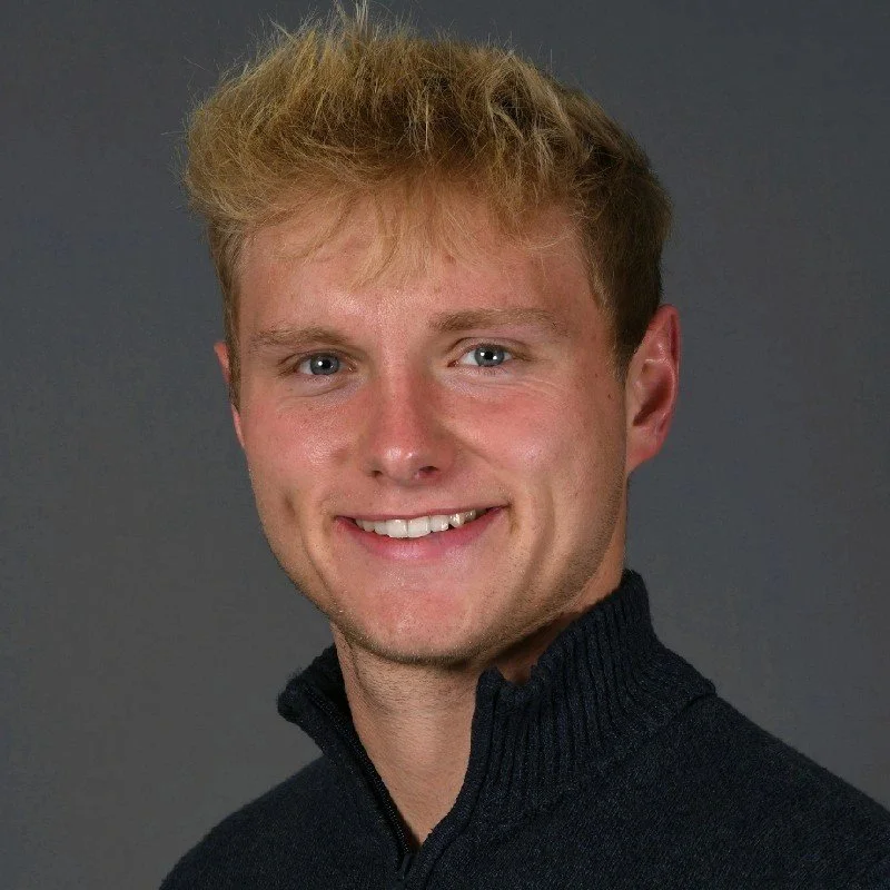 Portrait of a young man with blonde hair, blue eyes, and a friendly smile, wearing a black sweater with a zip collar.