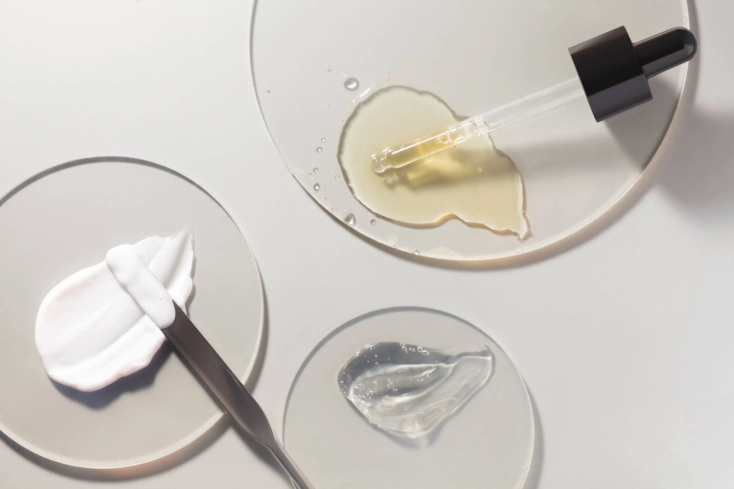 A dropper releasing a clear liquid into petri dishes, some containing a creamy substance and others empty, on a white surface.