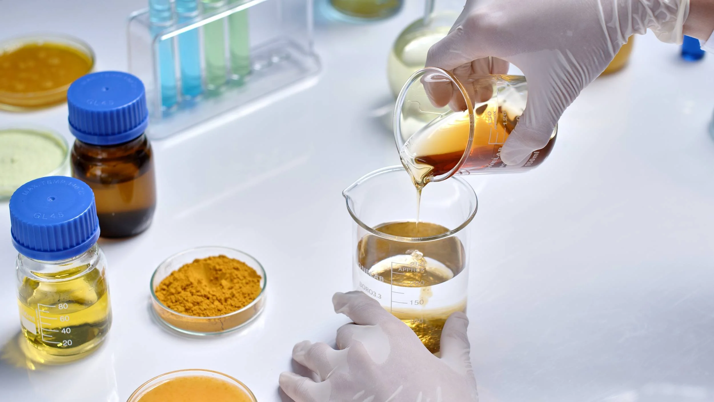 A person wearing gloves pouring a yellowish liquid from a test tube into a beaker in a laboratory setting. There are various laboratory bottles and petri dishes with yellow and orange substances on the white surface.