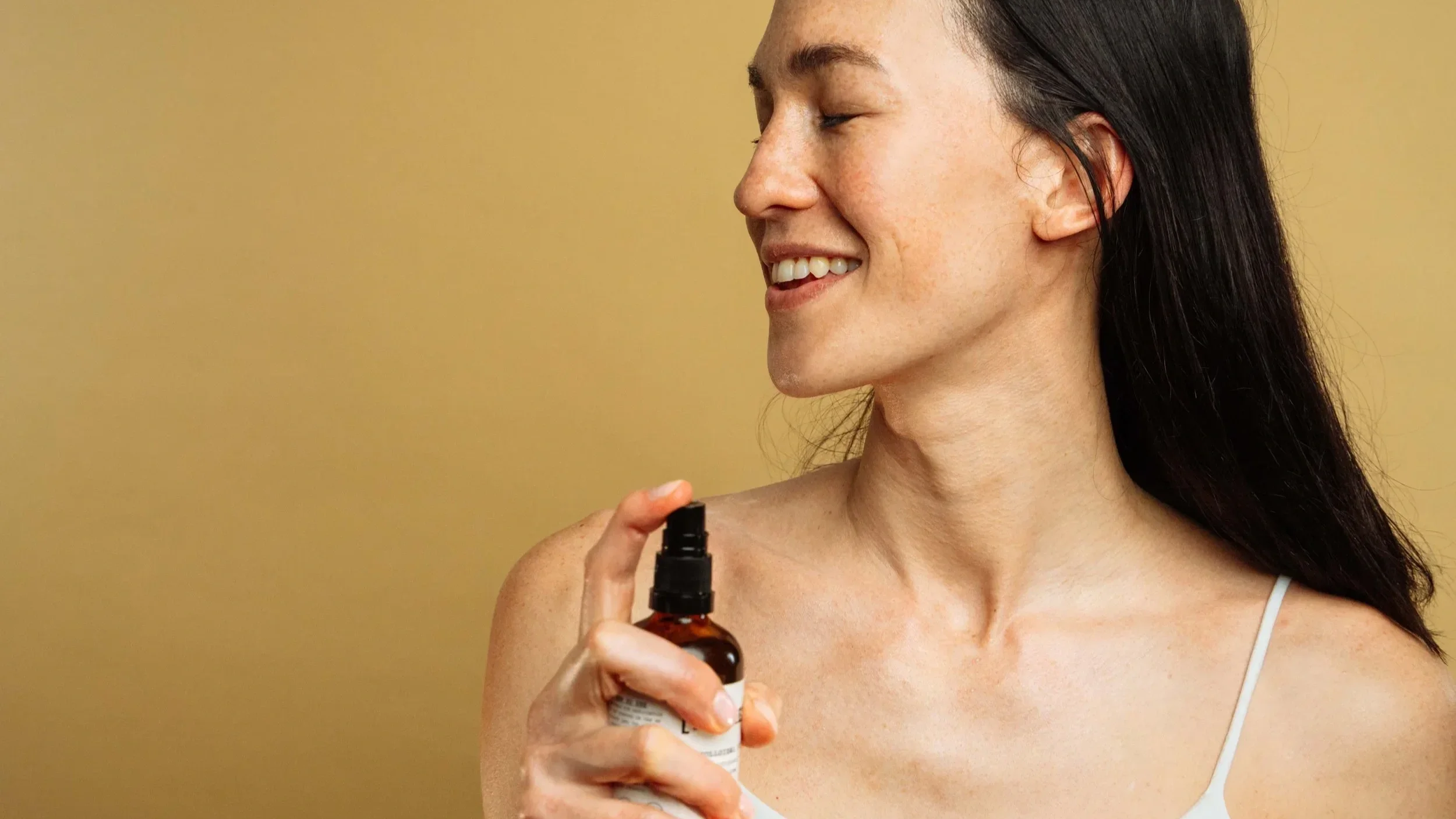 A young woman with long dark hair, smiling with eyes closed, holding a spray bottle in one hand.
