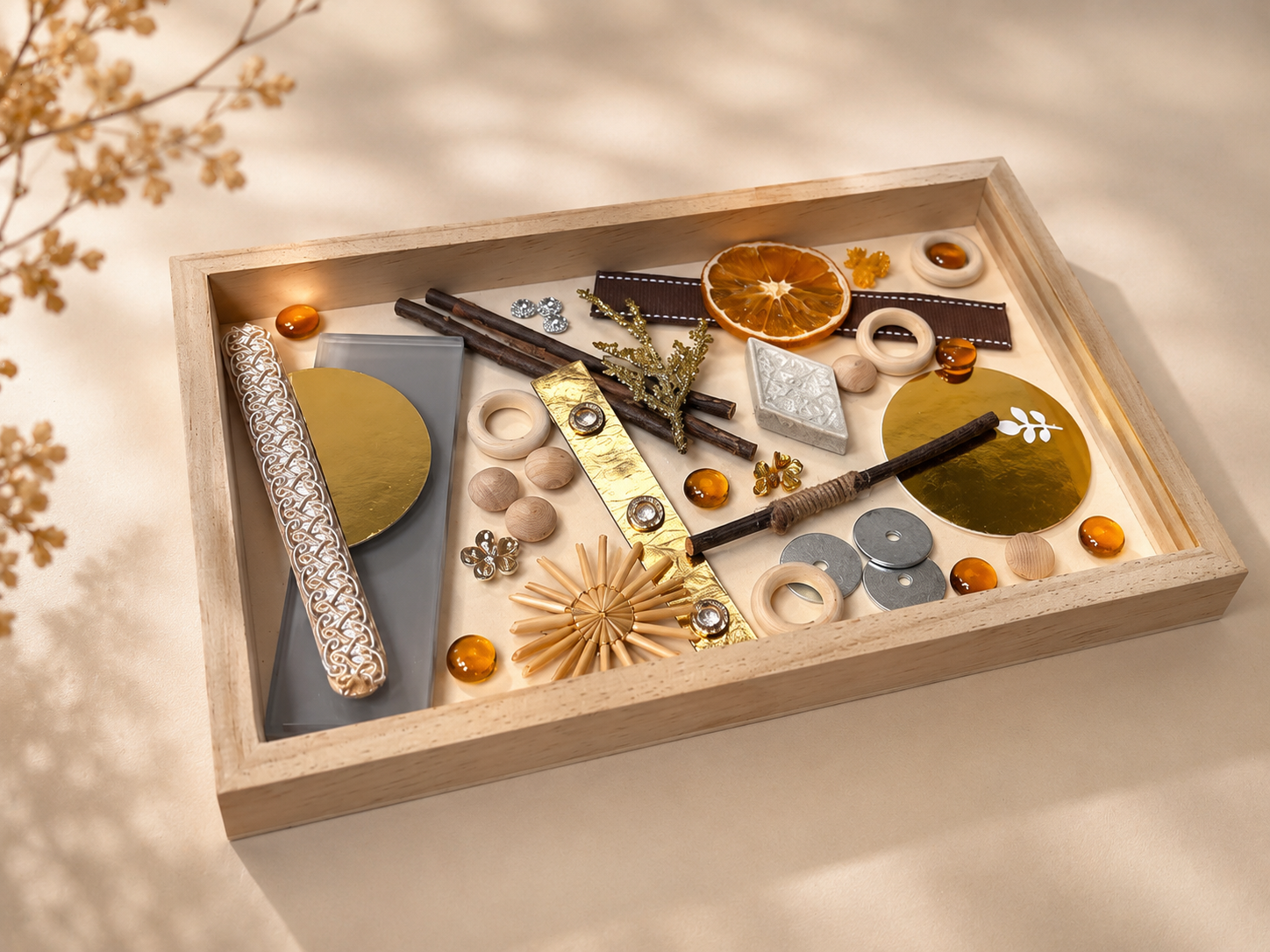 A wooden tray containing decorative items such as dried orange slices, cinnamon sticks, metallic and wooden embellishments, and glass beads, arranged on a beige surface.