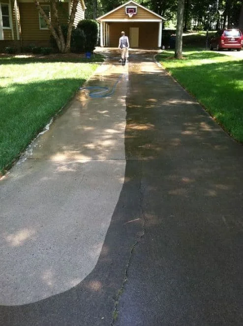 A concrete driveway partially cleaned, with the cleaned section on the left and the uncleaned, darker section on the right. A person is in the background, standing near the garage, which has a basketball hoop above it. The driveway is flanked by green grass and trees.