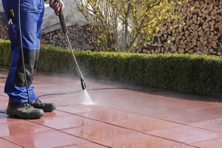 Person power washing a red concrete patio with a high-pressure spray, wearing work boots and blue work pants, with trimmed bushes and a woodpile in the background.