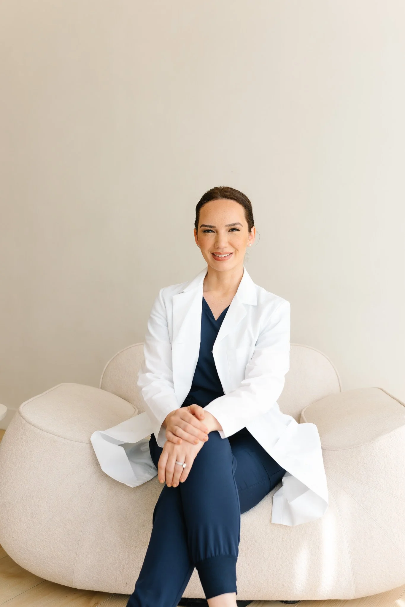 A woman sitting on a curved beige sofa, smiling, wearing a white lab coat over navy blue scrubs, with dark hair pulled back, in a neutral-colored room.
