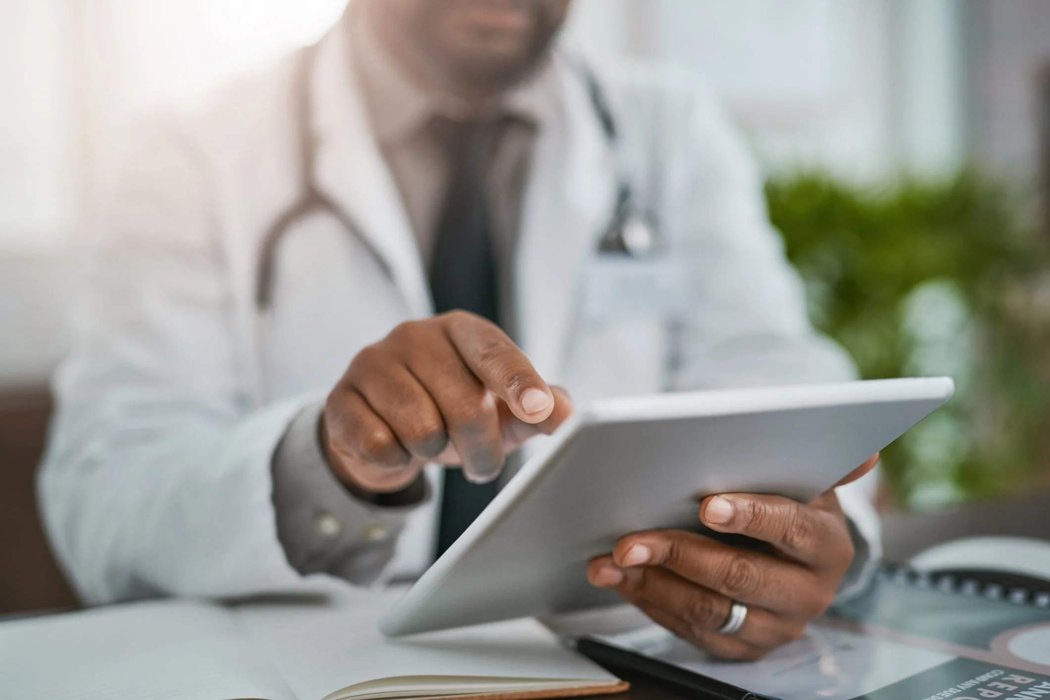 Close-up of a man wearing a white coat and black tie, holding a digital tablet, sitting at a table with a notebook and a pen. The background is blurred, featuring greenery and a window, suggesting an indoor setting.