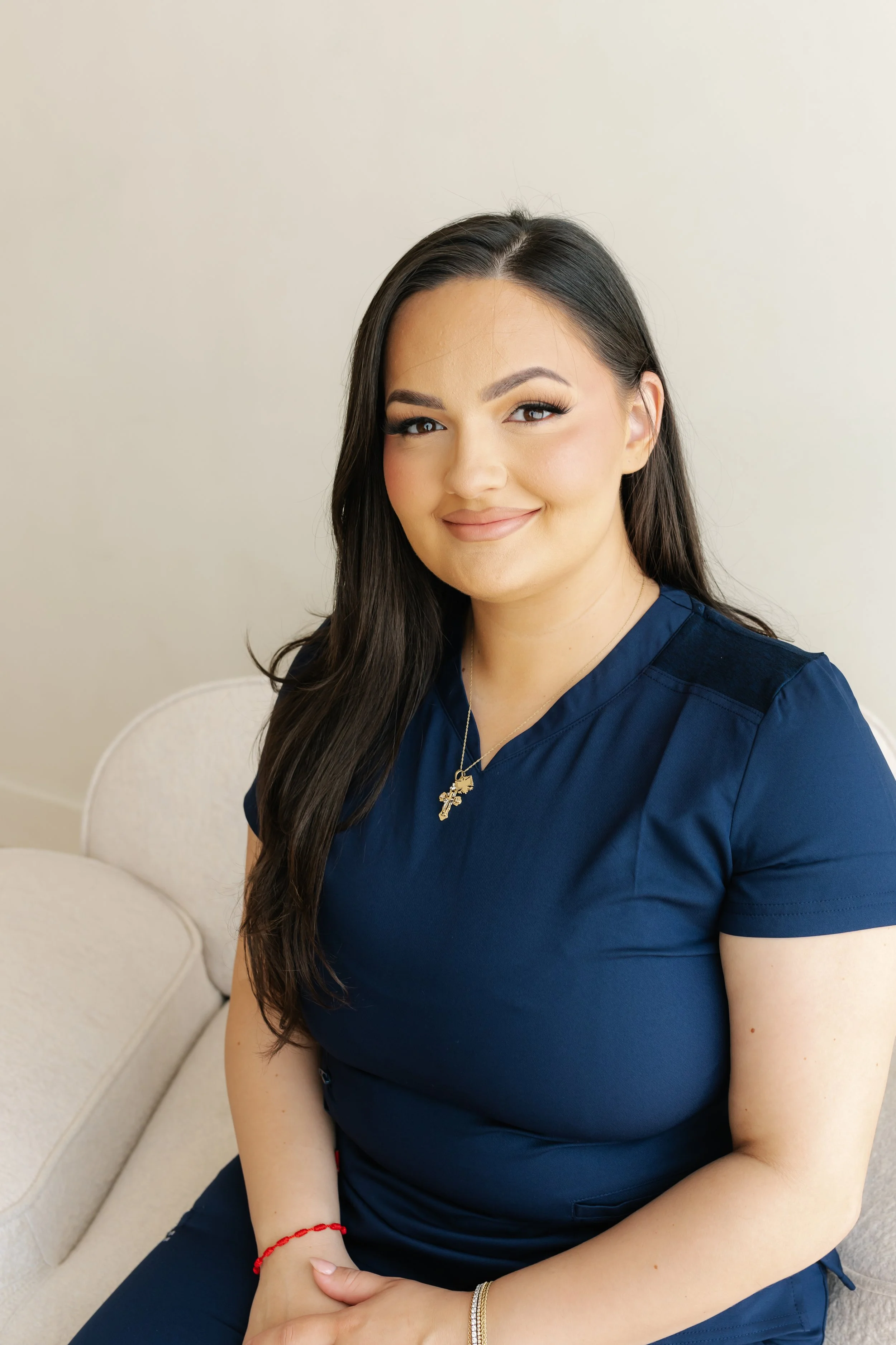 A young woman with long dark hair, wearing a dark blue scrub top and jewelry, smiling with her eyes closed, sitting on a cream-colored chair against a plain light-colored wall.
