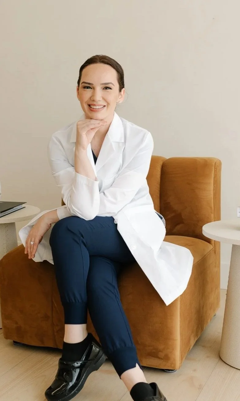 A woman smiling and sitting on a brown chair, wearing a white lab coat, navy pants, and black shoes, in a room with a beige wall and a small white side table.